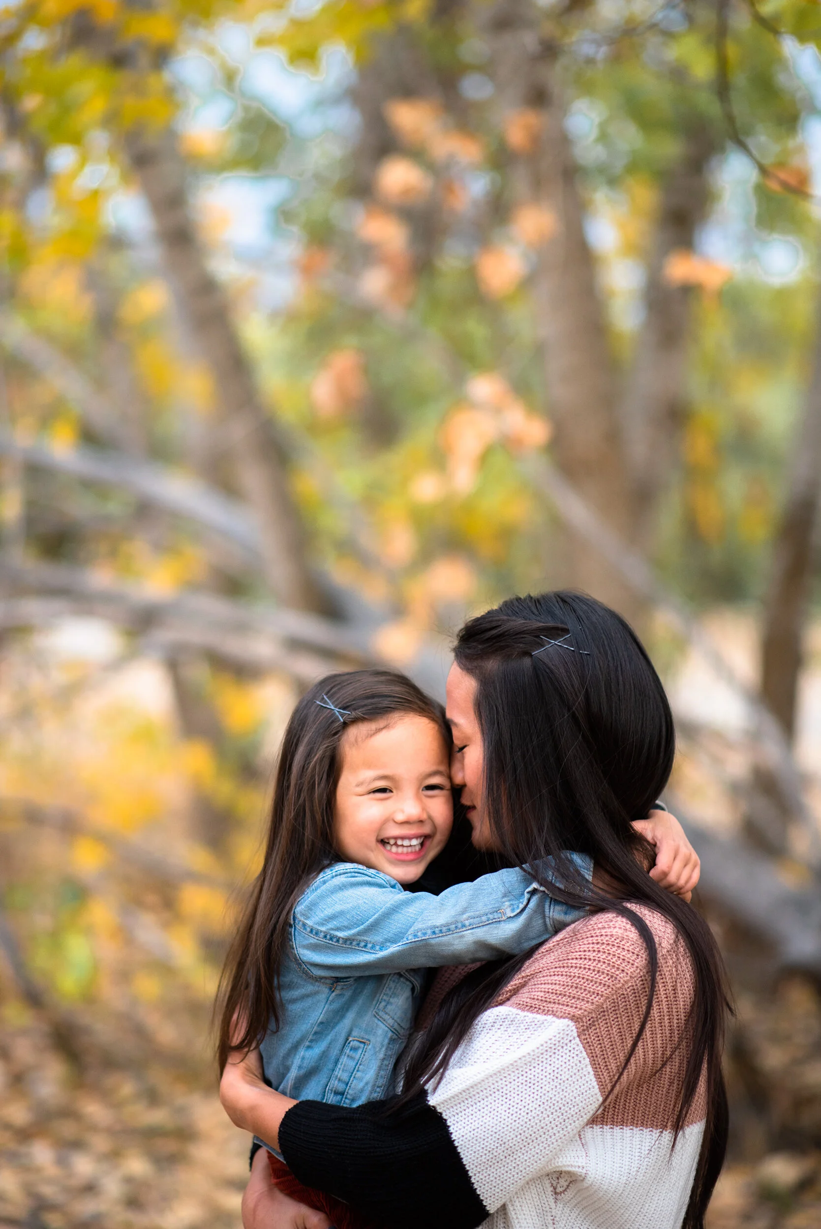 A woman and a young girl hugging outdoors, surrounded by autumn-colored trees.