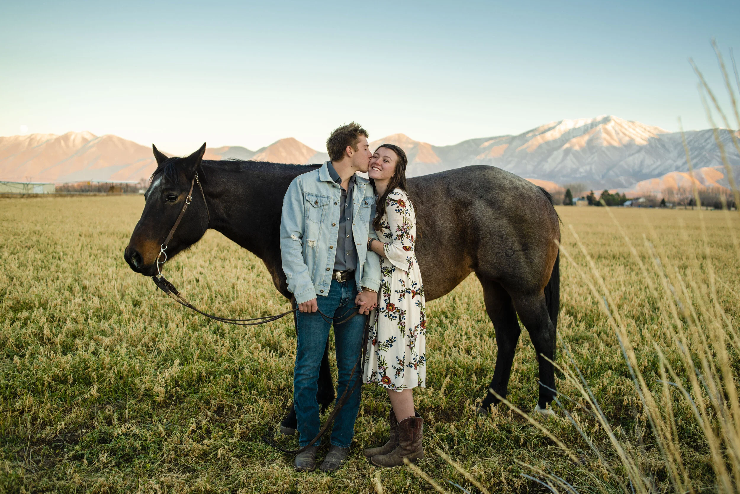 A couple stands in a grassy field with a horse, holding hands. The man is kissing the woman's cheek, both smiling, with snow-capped mountains in the background.