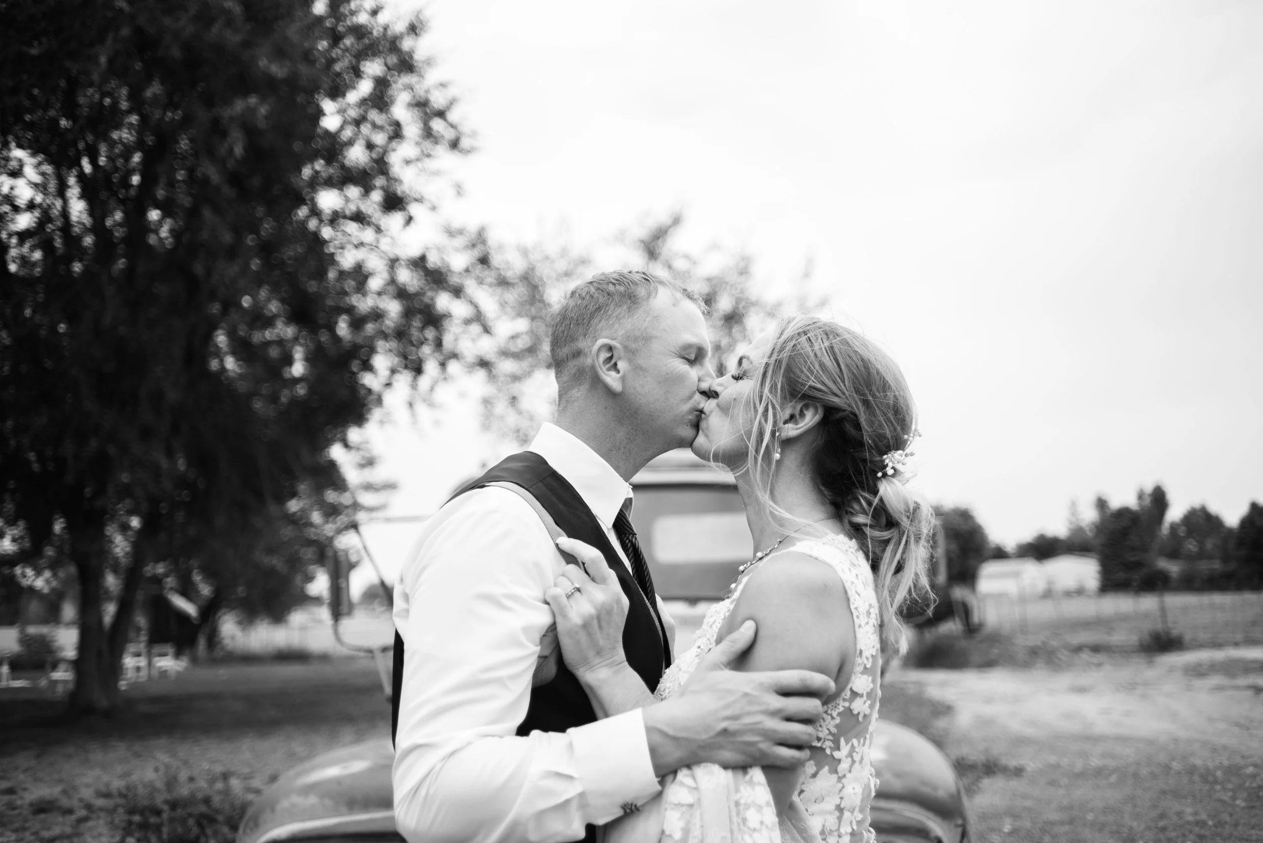 A black-and-white photo of a newlywed couple sharing a kiss outdoors, with trees and a vehicle in the background.