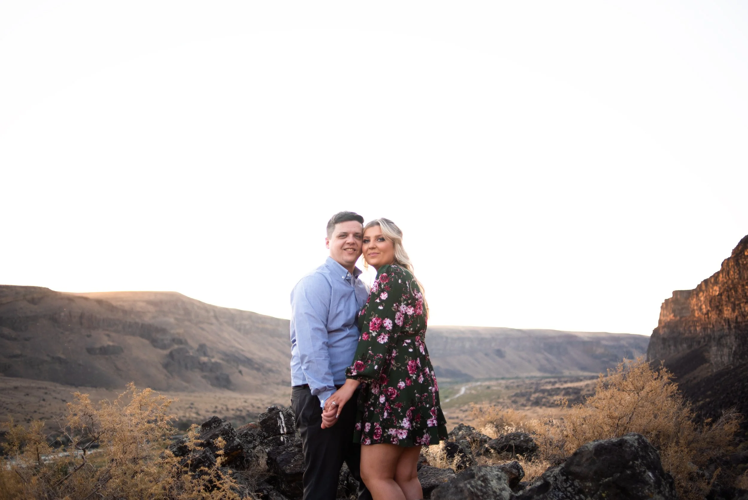 A couple standing together outdoors in front of a mountainous landscape, holding hands and looking at the camera during sunset.