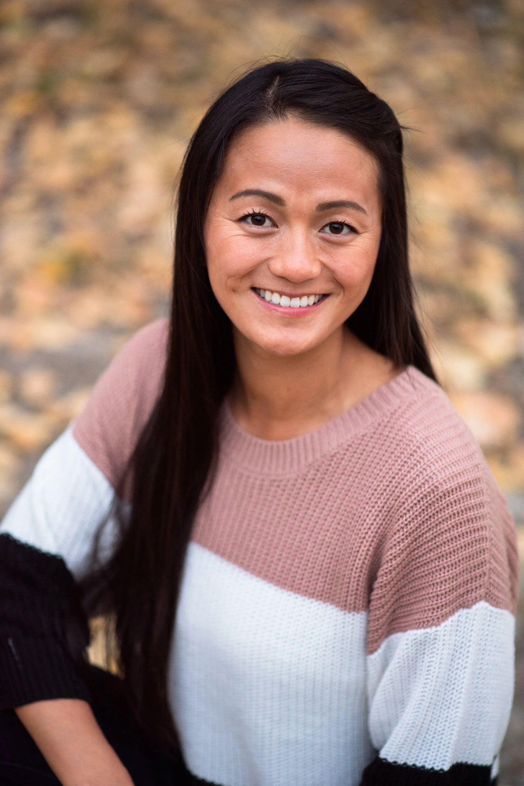 A smiling woman with long dark hair, wearing a color-blocked sweater in pink, white, and black, standing outdoors with a blurred background of autumn leaves.