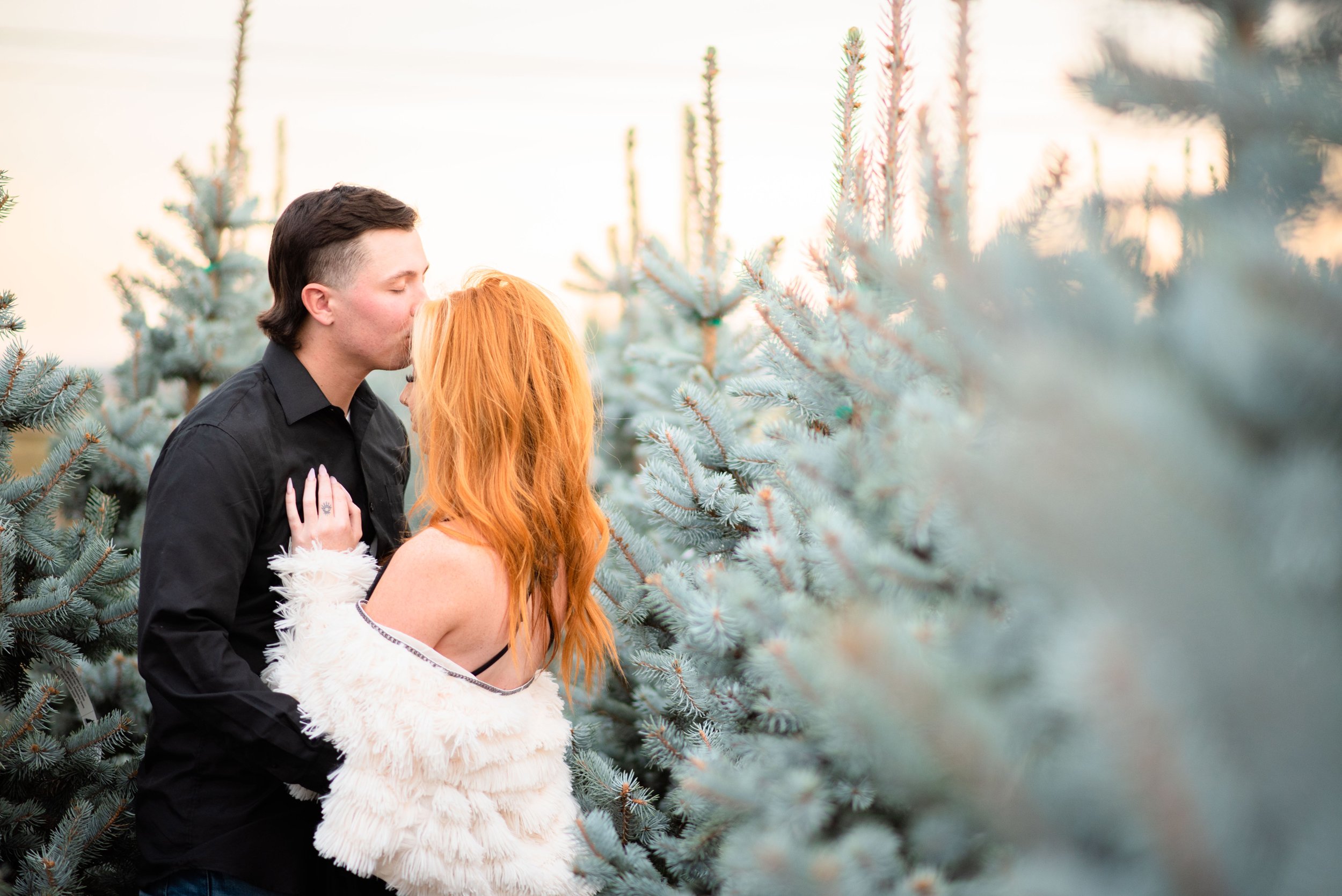 A man with dark hair and a woman with long red hair sharing a kiss among Christmas trees.