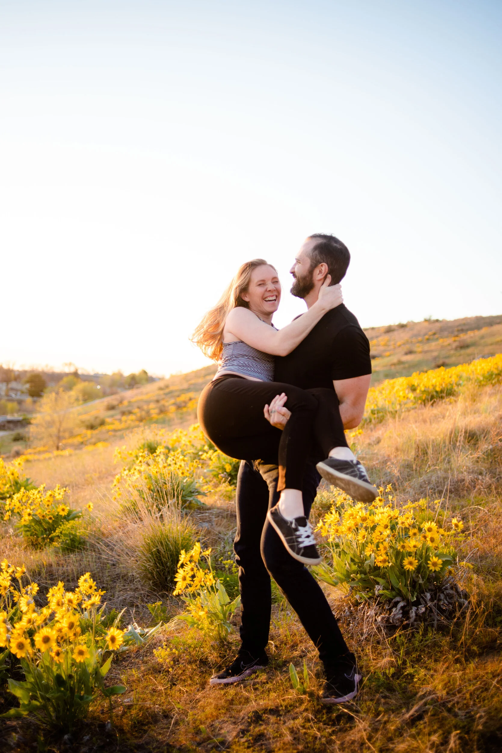 A man is carrying a woman in a field of yellow flowers during sunset, both are smiling and looking at each other.