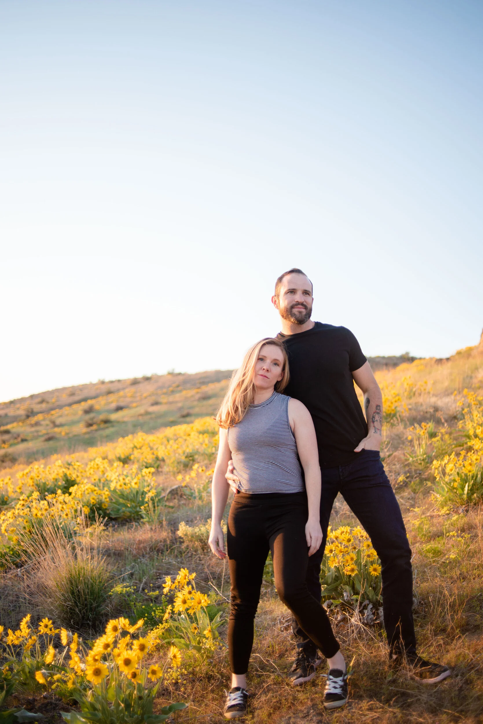 A man and a woman standing outdoors in a field of yellow flowers with a hill in the background during sunset.