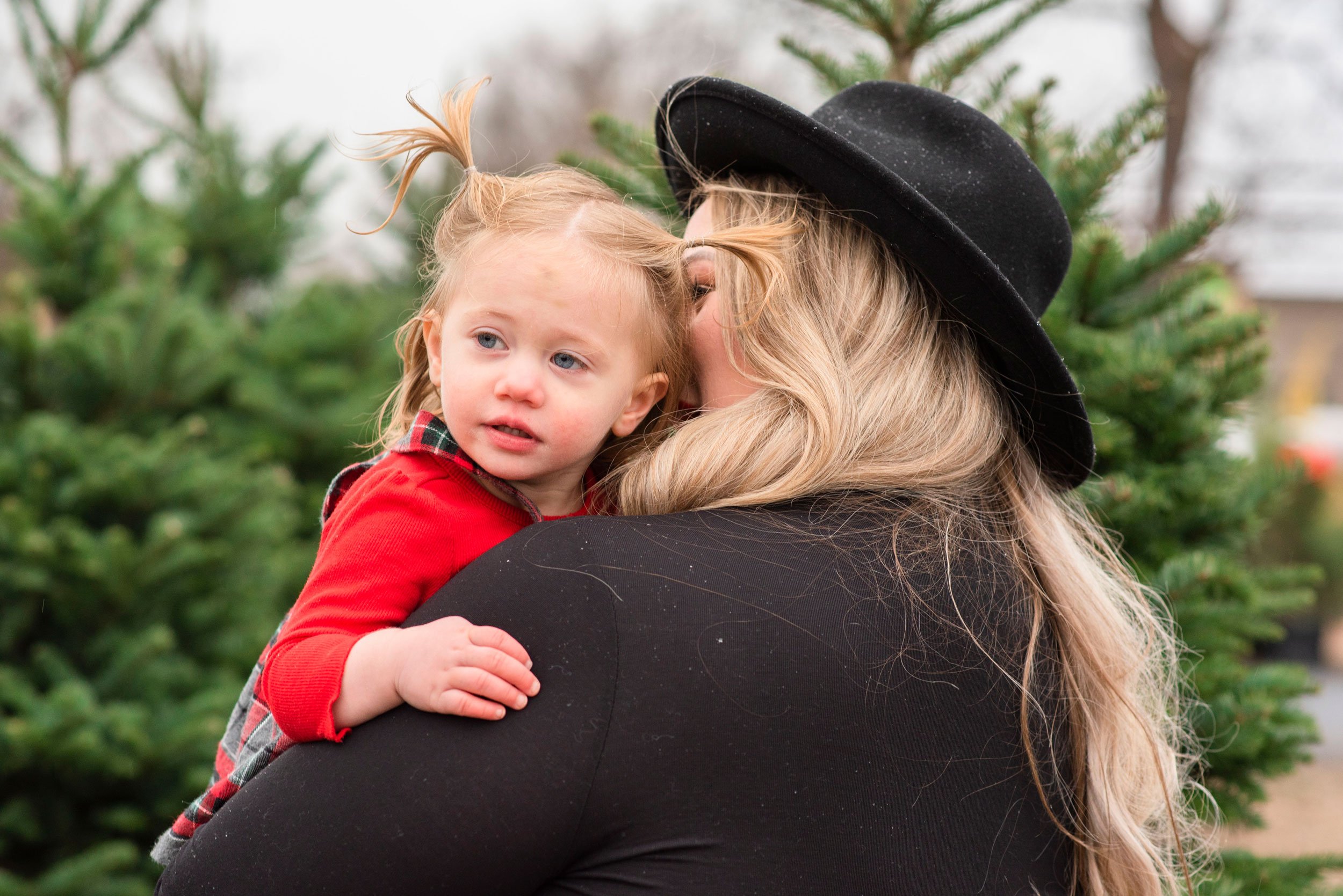 A woman wearing a black wide-brim hat holds a young girl with red hair in pigtails, outdoors surrounded by evergreen trees.