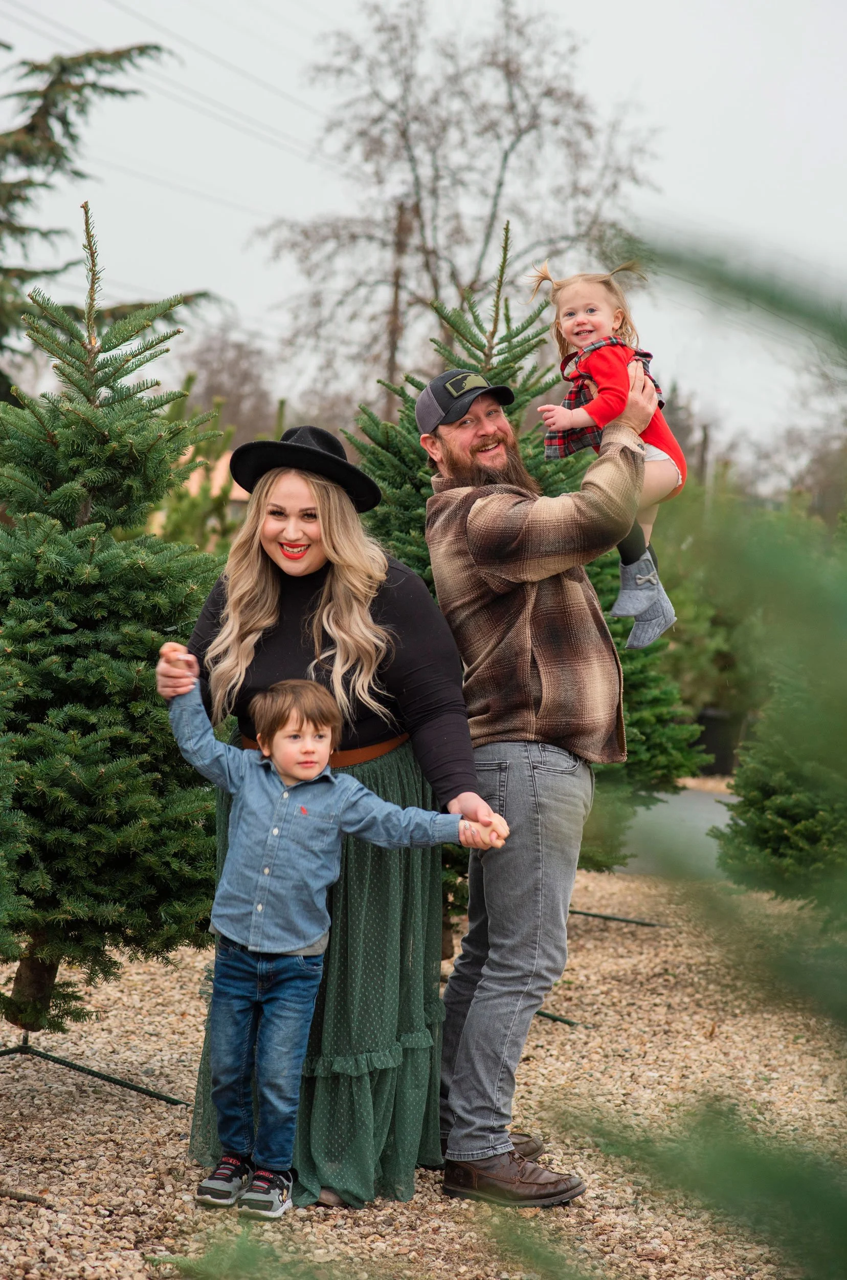 A family of four on a Christmas tree farm, surrounded by evergreen trees. The father is lifting a young girl in a red dress, while the mother and a young boy hold hands and smile at the camera. The scene is outdoors on a cloudy day with a gravel path