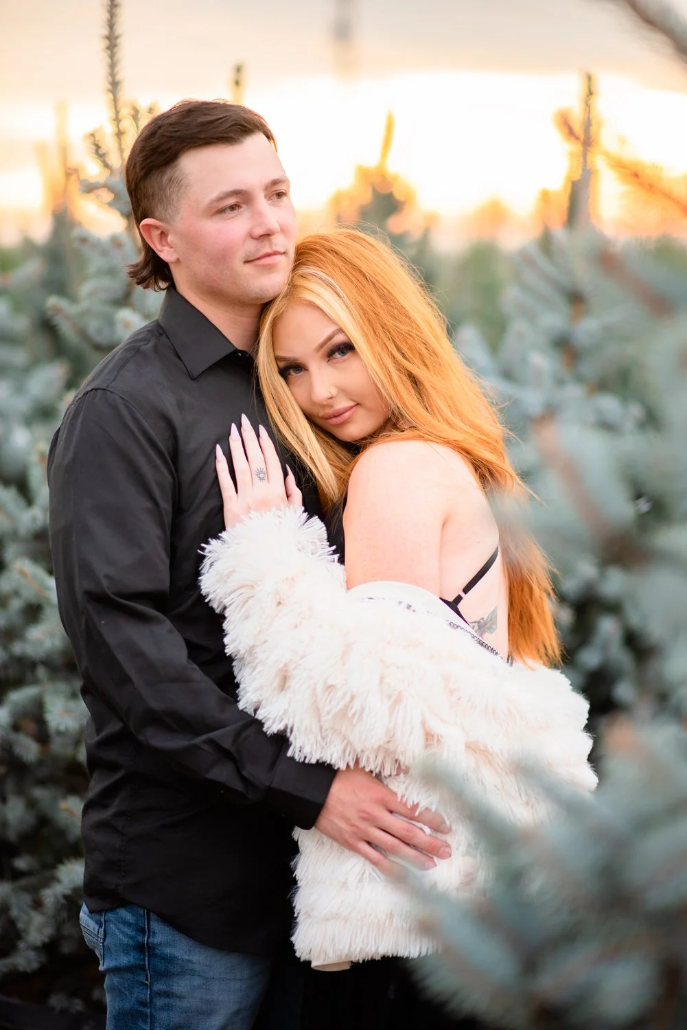 A young couple standing close together outdoors during sunset, with evergreen trees in the background. The man has short dark hair and is wearing a black shirt, while the woman has long red hair and is wearing a white furry jacket and black top. She 