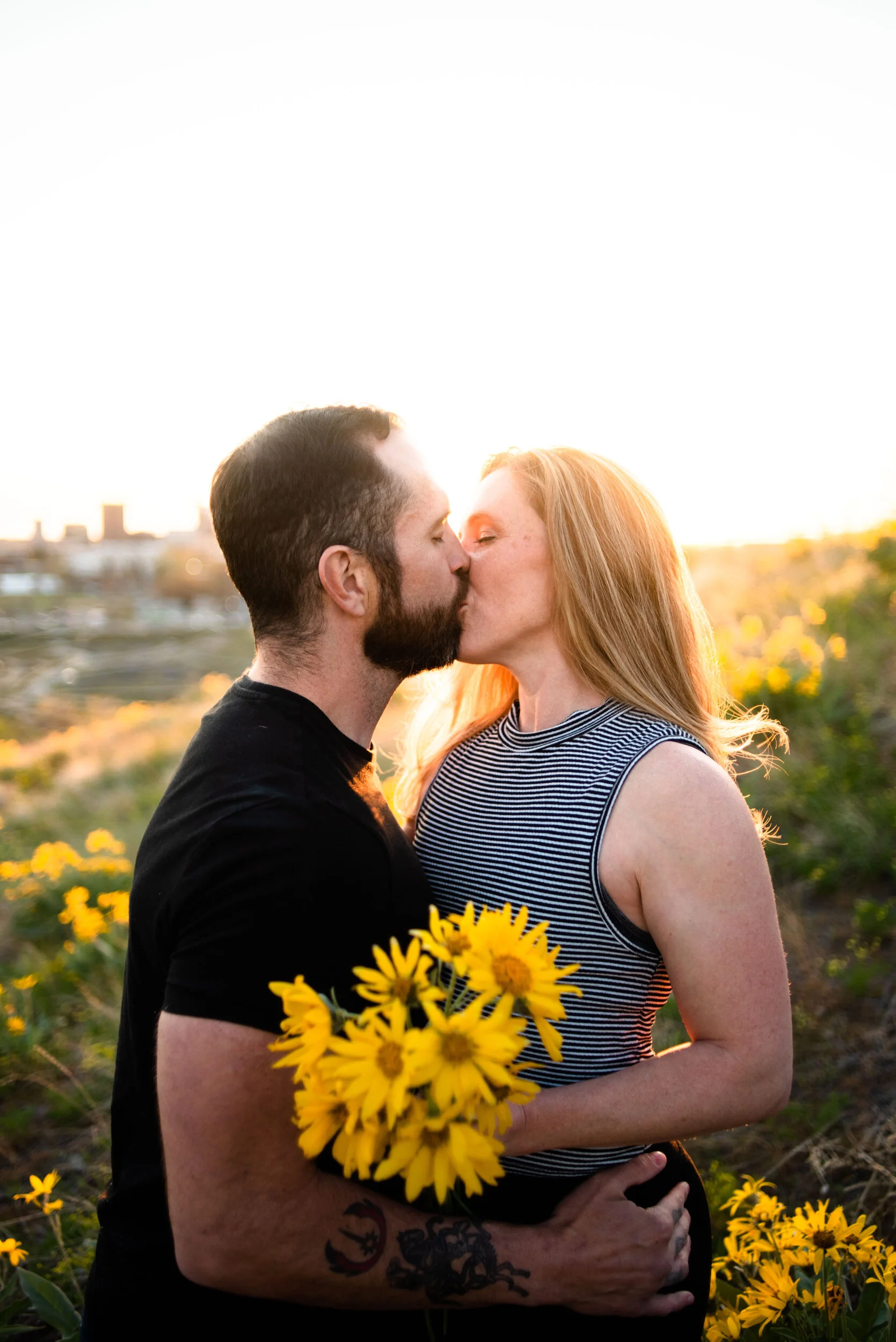A couple is kissing in a field of yellow flowers at sunset, with the man holding a bouquet of yellow flowers.