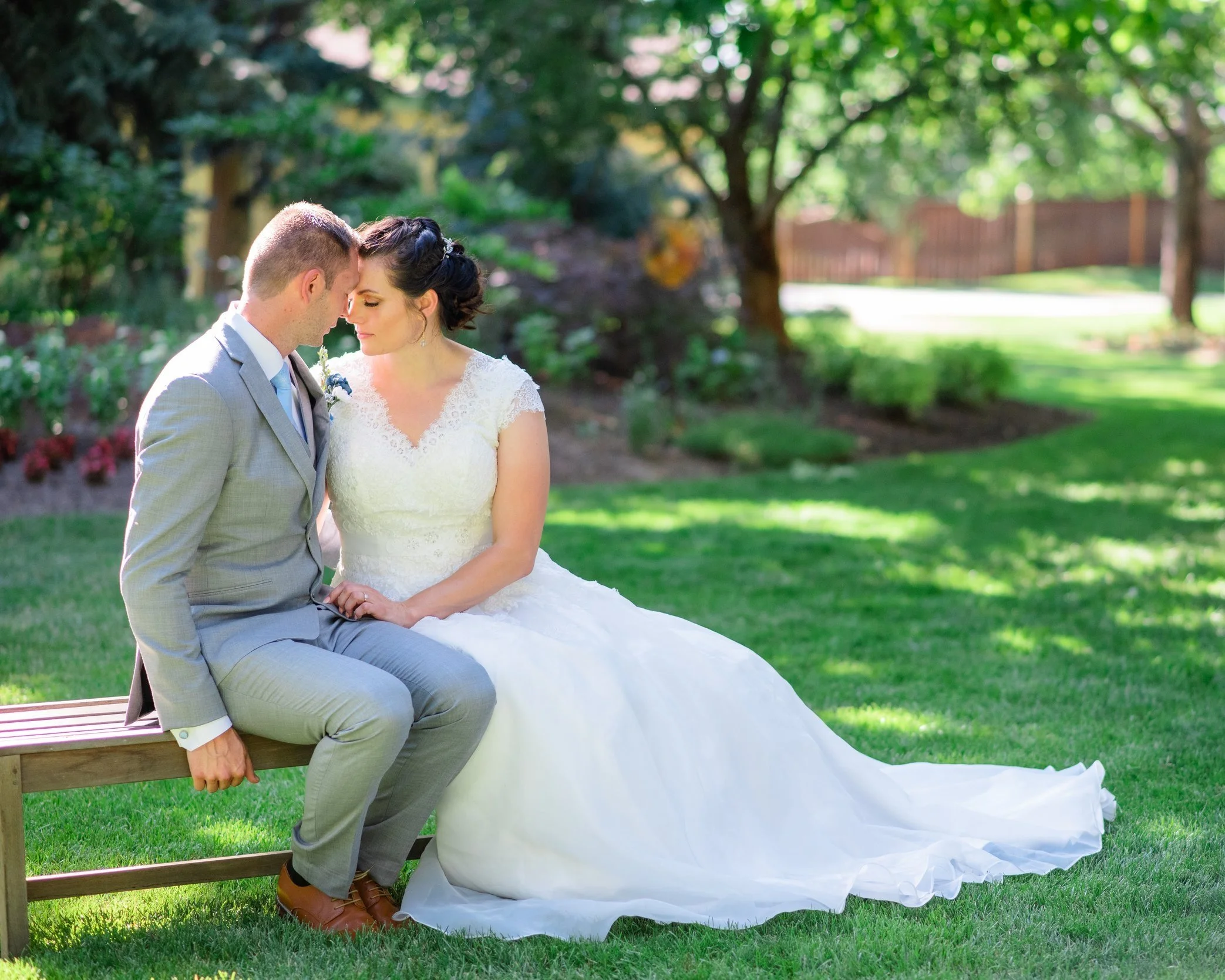 A bride and groom sitting closely together on a park bench with their foreheads touching, outdoors in a lush green garden during daytime. Idaho Wedding Photographer, Burley
