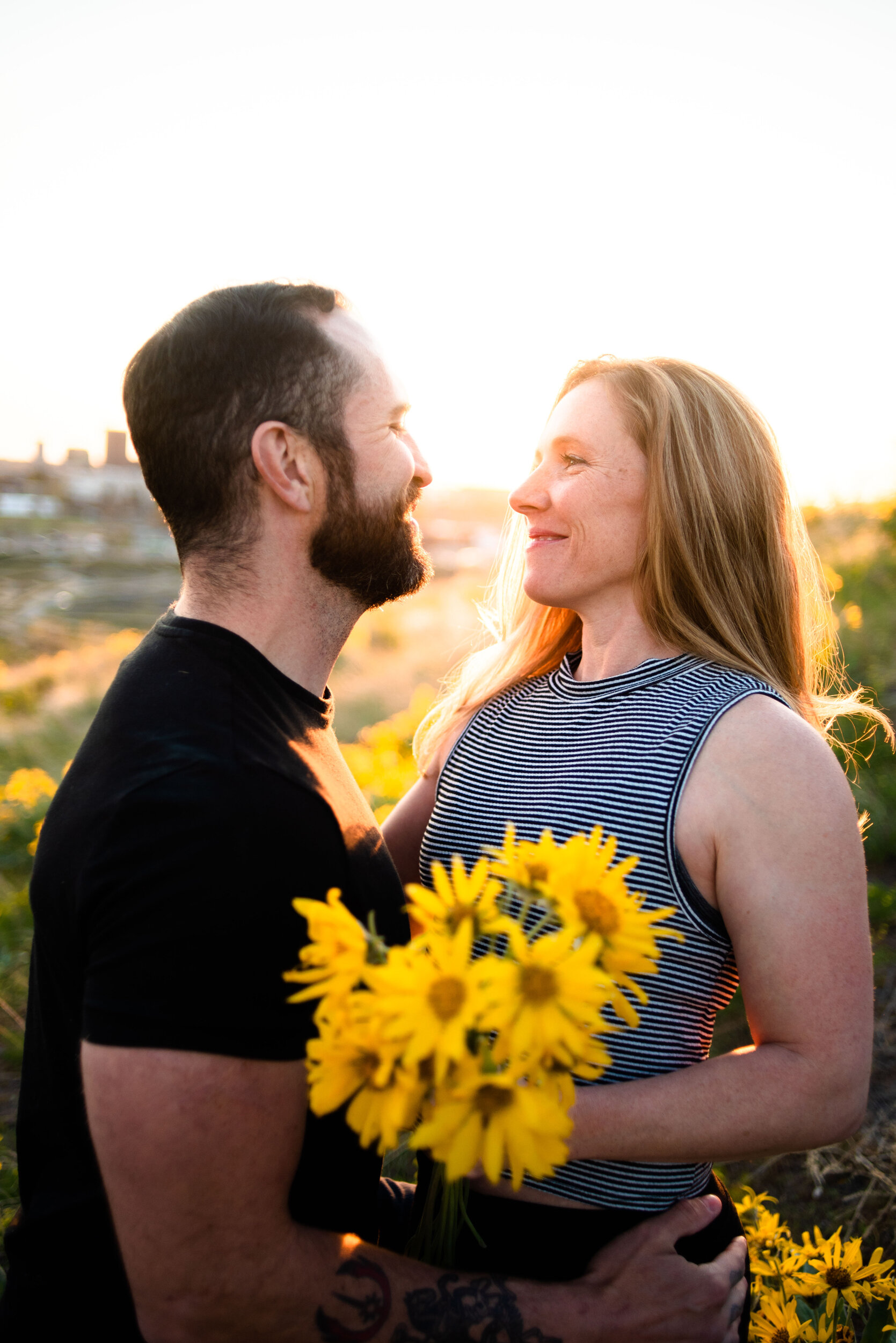 A couple sharing a close, joyful moment outdoors during sunset, with the woman holding a bouquet of yellow flowers.