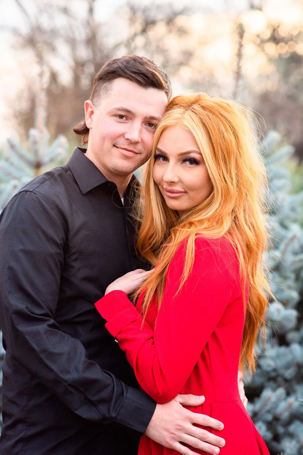 A young couple embracing outdoors, the man wearing a black shirt and the woman in a red dress, with trees and shrubs in the background.