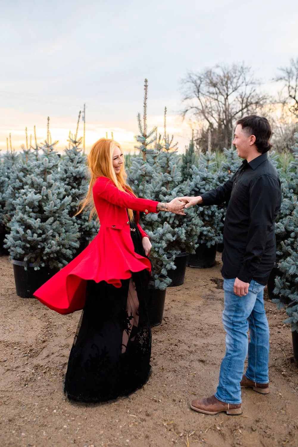 A woman in a red coat and black dress smiling and holding hands with a man in a black shirt and jeans, standing in a Christmas tree lot with blue spruce trees during sunset.