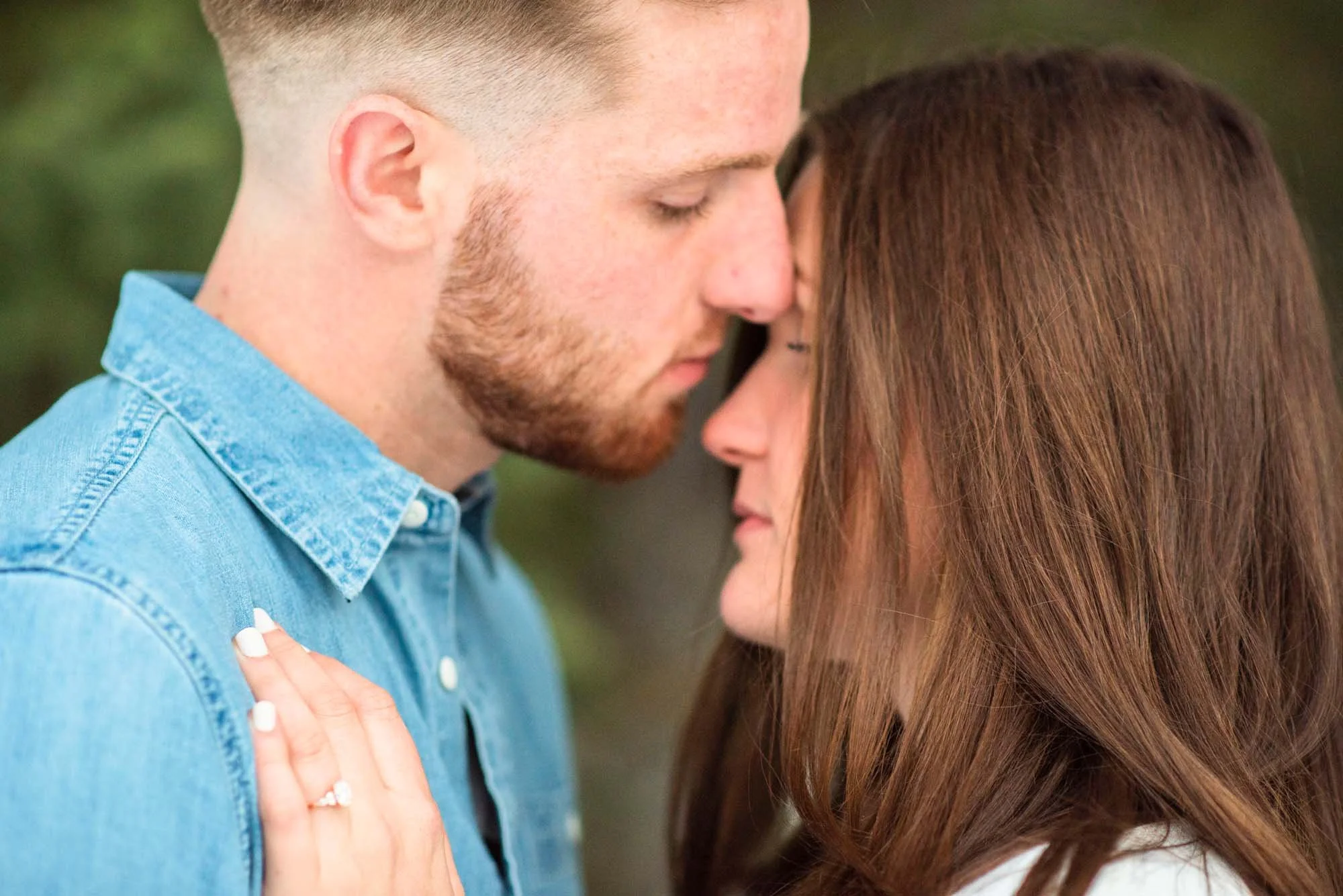 A couple with their foreheads touching, eyes closed, in a tender moment outside. The man has short light hair and a beard, wearing a light blue denim shirt. The woman has long reddish-brown hair with a hand resting on his chest.