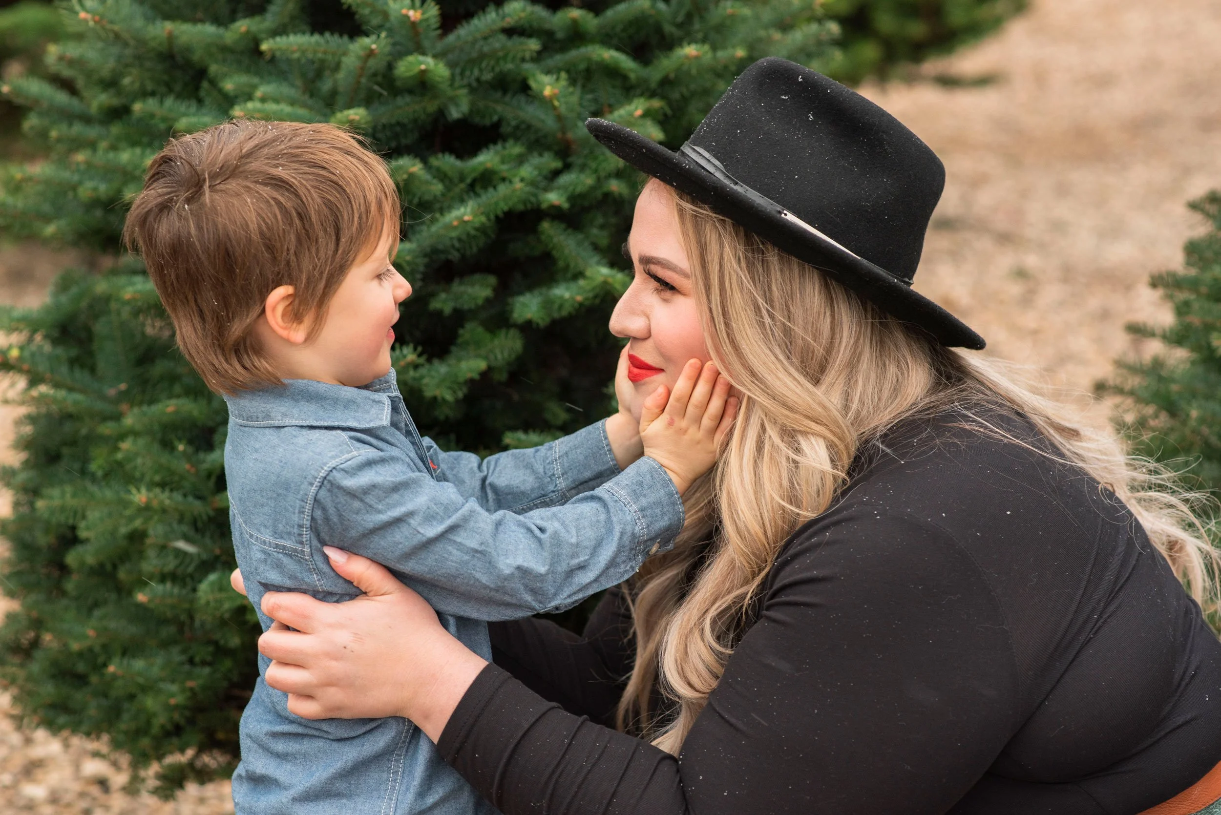 A young woman with long blonde hair, wearing a black hat and black coat, is gently holding a small boy's face, both smiling at each other near a Christmas tree outdoors.