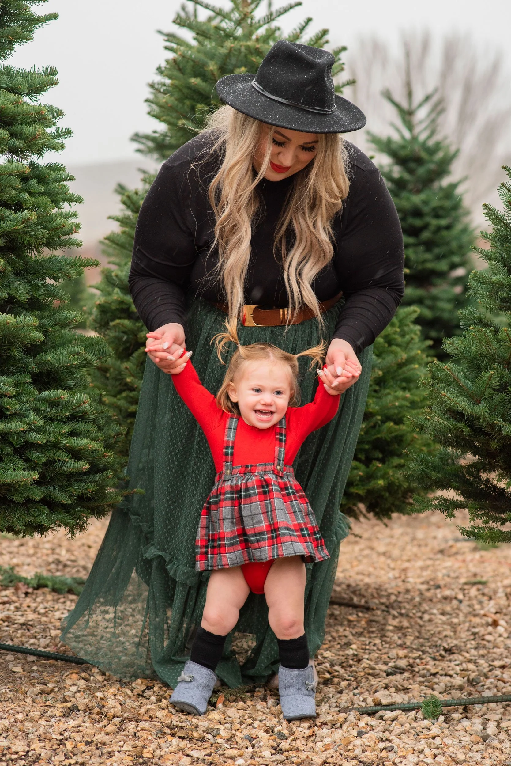 A woman with long blonde hair wearing a black hat and black sweater is holding hands with a young girl in a red and plaid dress, standing between Christmas trees outdoors.