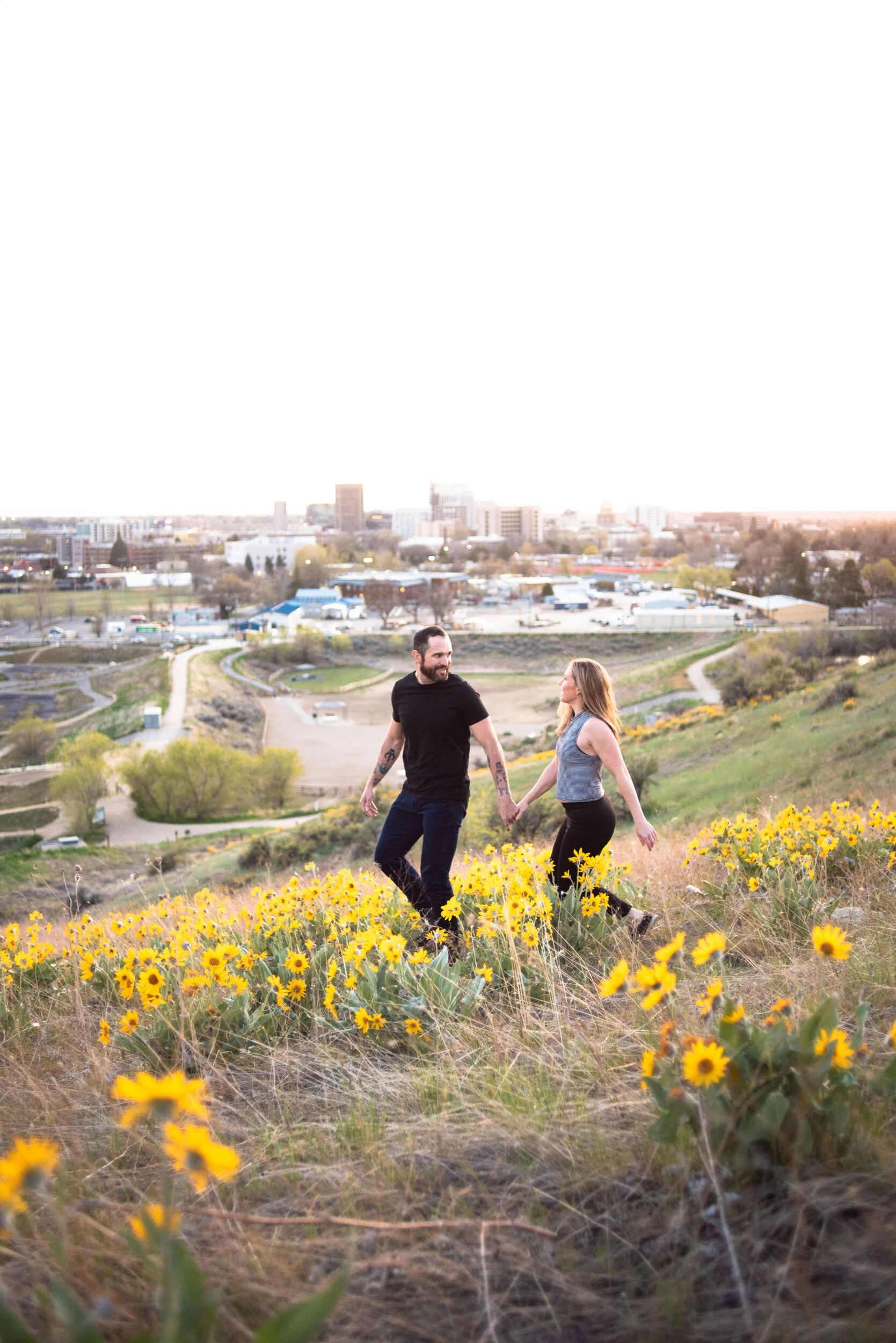 A couple holding hands walks through a field of yellow wildflowers on a hillside, with a cityscape in the background during sunset.