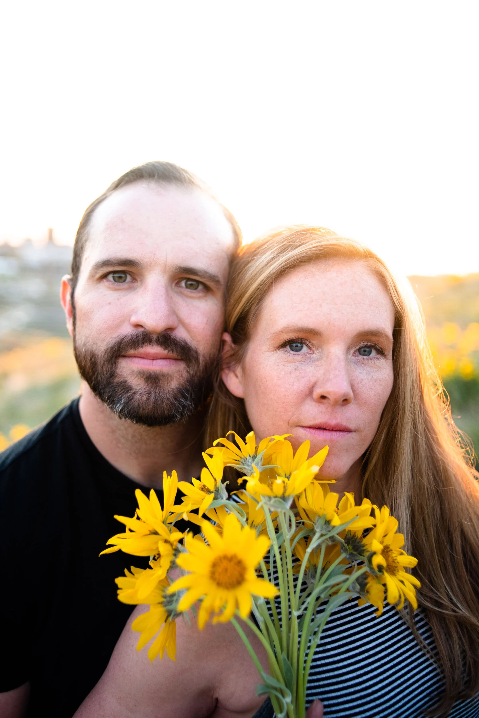 A man and a woman are close together, with the man on the left and the woman on the right, holding a bouquet of bright yellow sunflowers partially covering her chest. They are outdoors during sunset or sunrise, with blurred nature and cityscape in th