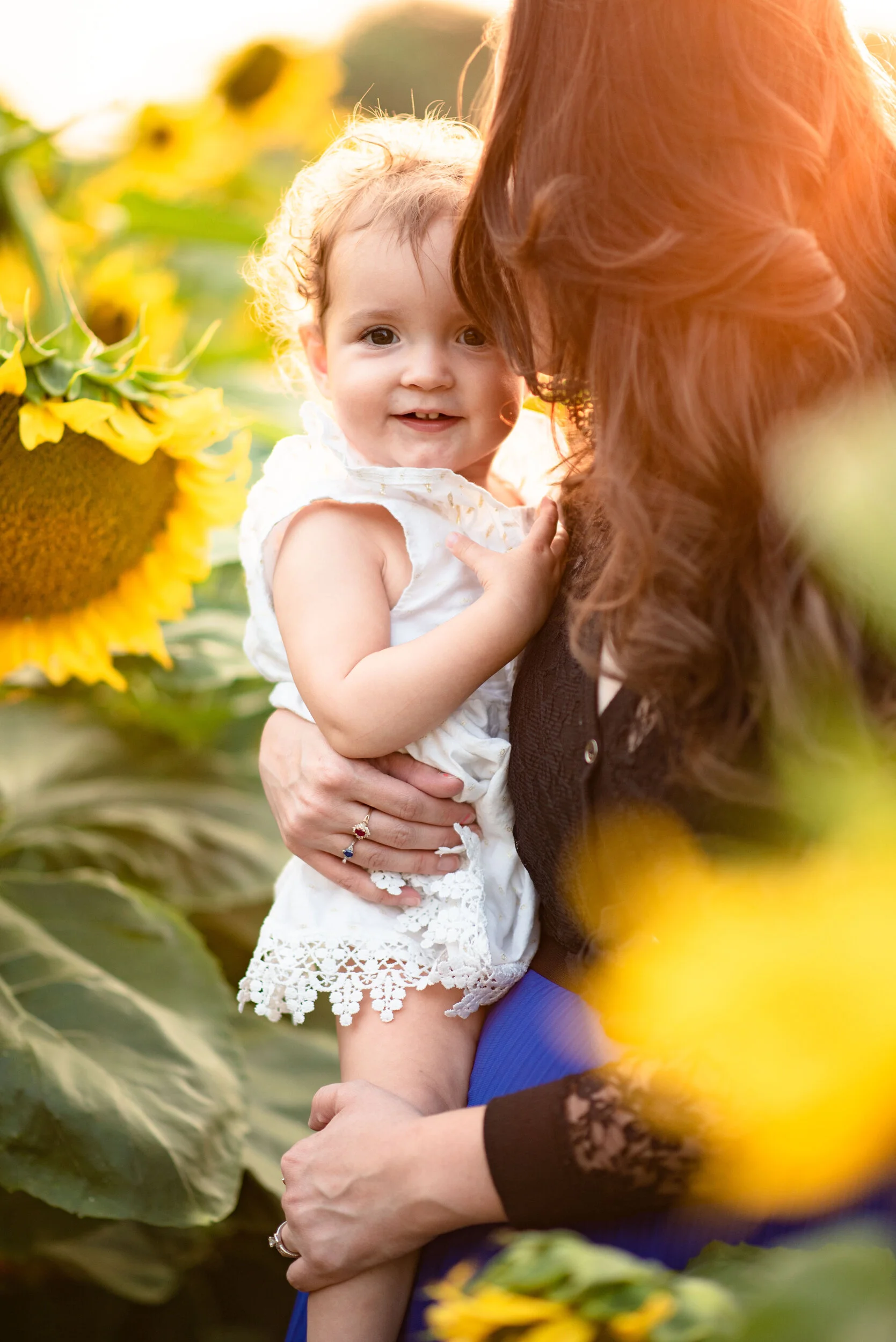 Close-up of a smiling toddler girl with curly hair being held by a woman, in a sunflower field during golden hour sunlight.