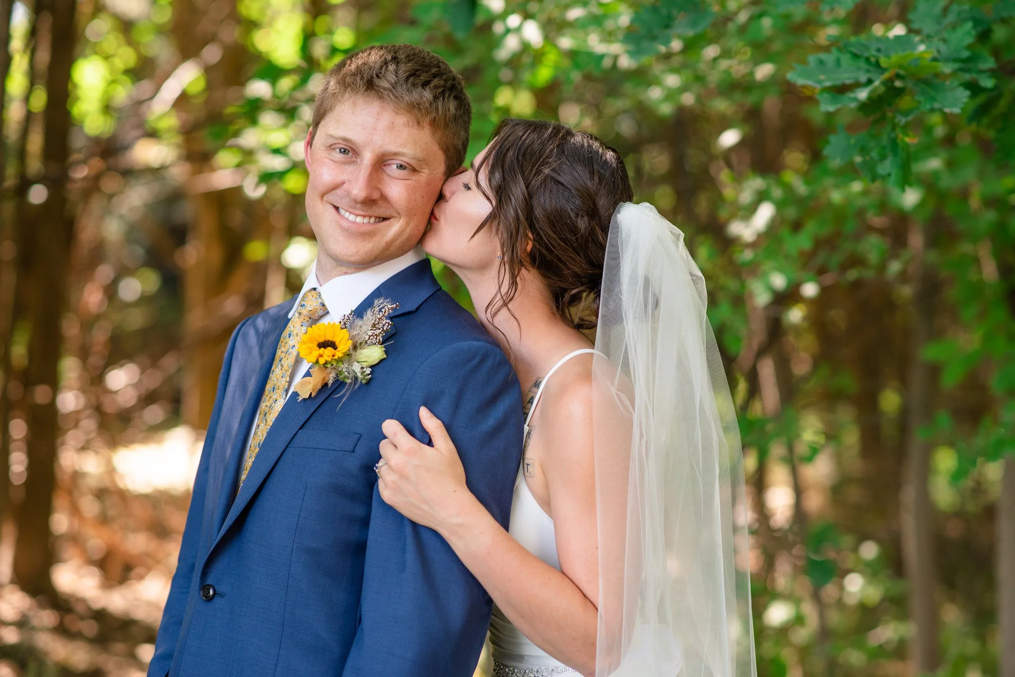 A newlywed couple sharing a kiss outdoors during daytime, surrounded by green trees in the background. The groom is wearing a blue suit with a yellow floral tie and a sunflower boutonniere, while the bride is wearing a white wedding dress and a veil.