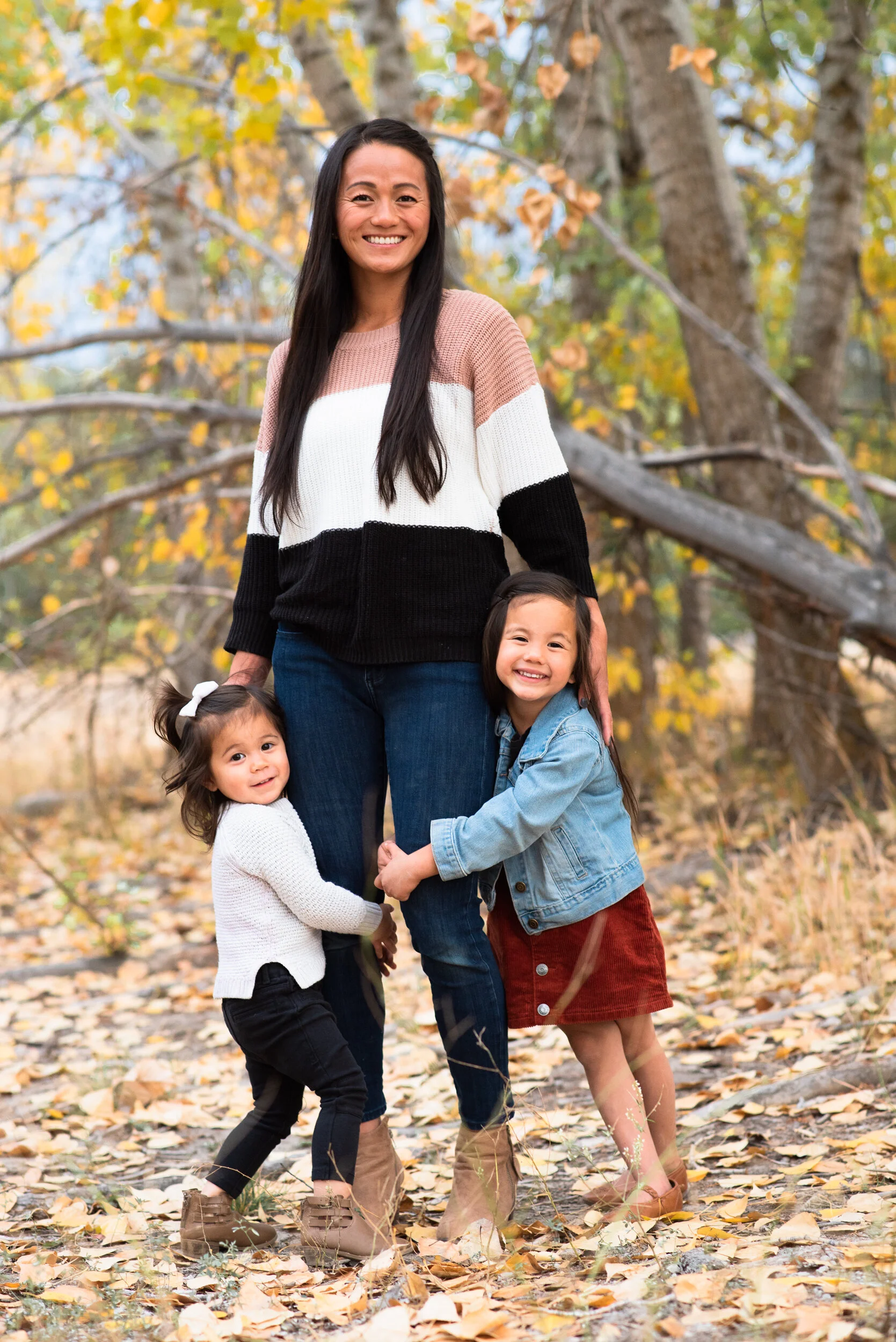 A woman and three young girls outside in autumn, standing on a leaf-covered trail, surrounded by trees with yellow and brown leaves. The woman is smiling and has long dark hair, wearing a color-blocked sweater, and the girls are hugging her, smiling 