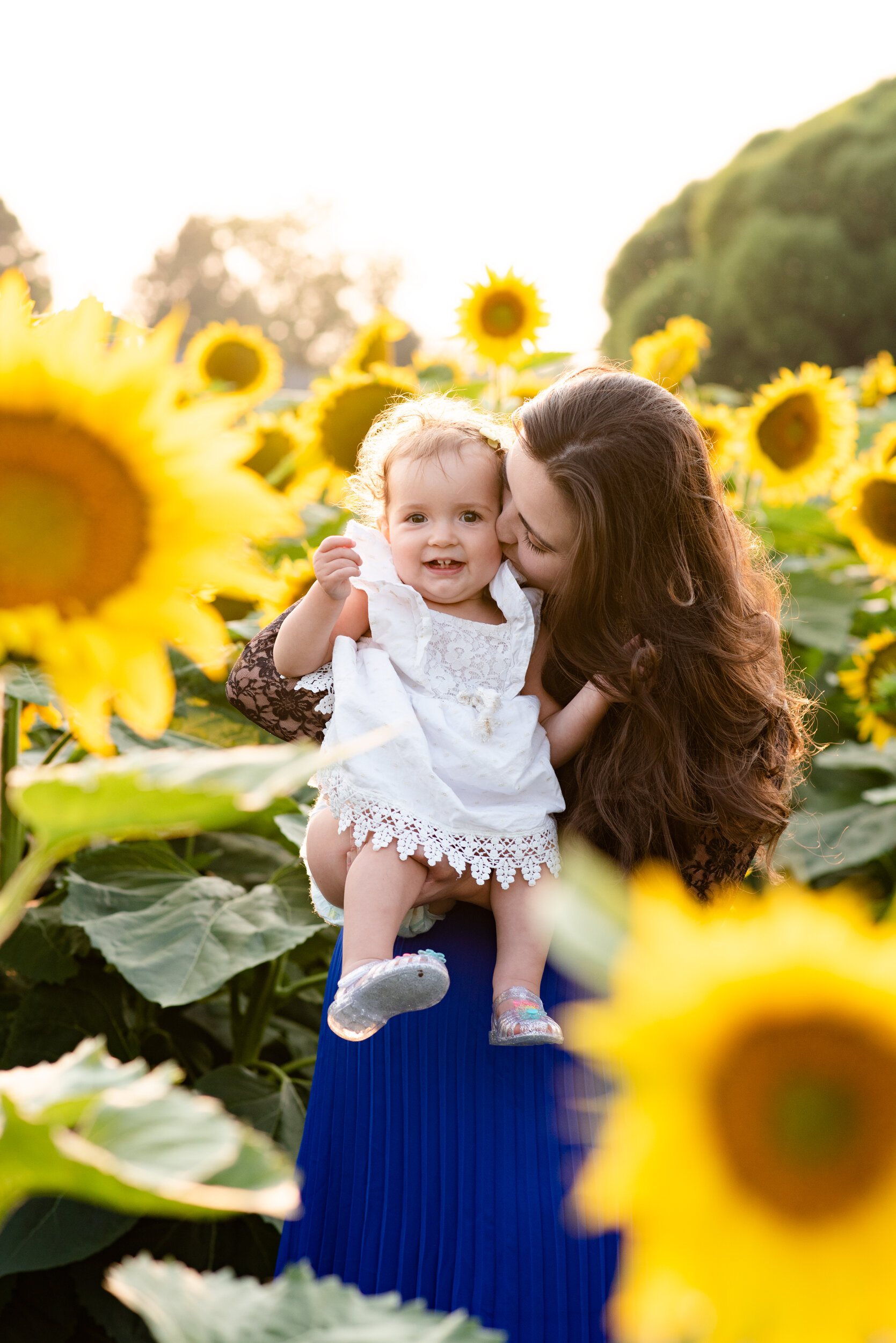 A woman with long brown hair and a blue skirt holds a smiling young girl with curly hair in a white dress among yellow sunflowers during sunset.