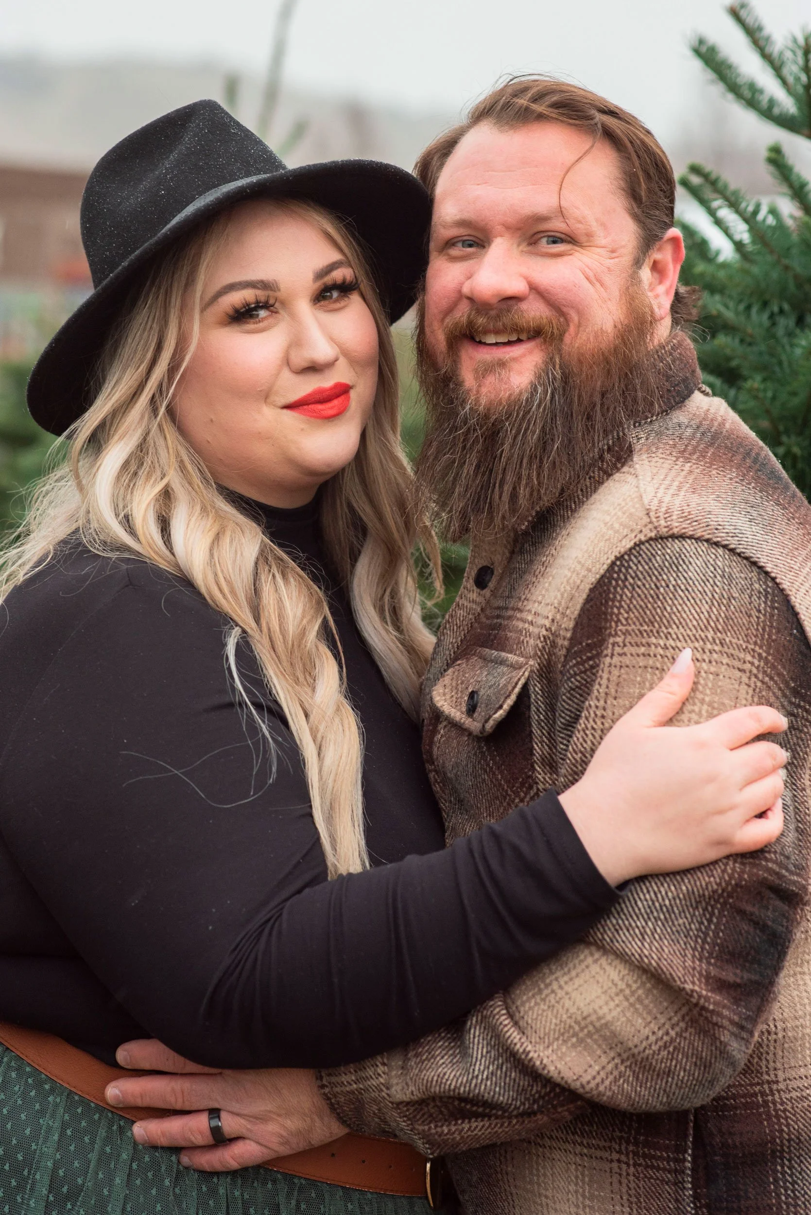 A woman with blonde hair, wearing a black wide-brimmed hat and black top, and a man with brown hair and a beard, dressed in a brown plaid jacket, are standing close together outdoors. They are smiling, with the woman embracing the man, who also has h