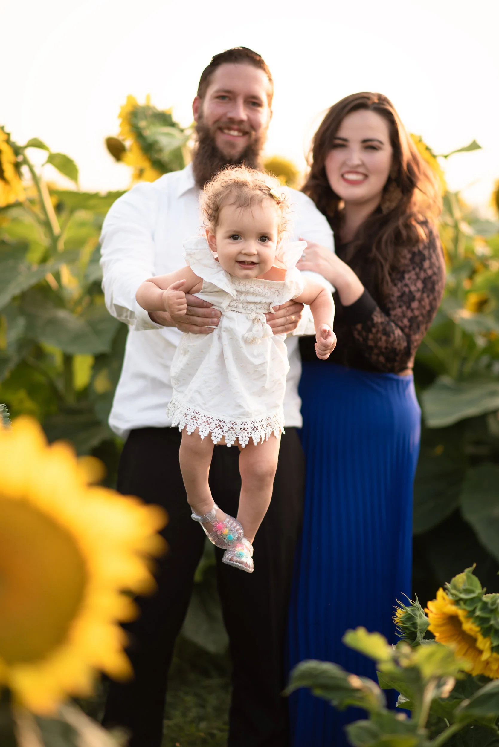 A family of three smiling and posing in a field of sunflowers, with the father holding the toddler girl, the mother standing next to them, all enjoying a sunny day.