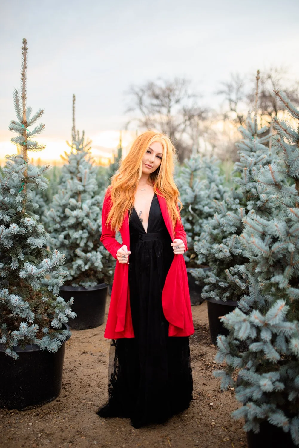 A woman with long red hair standing among potted blue spruce trees during sunset, wearing a black dress and a red coat.
