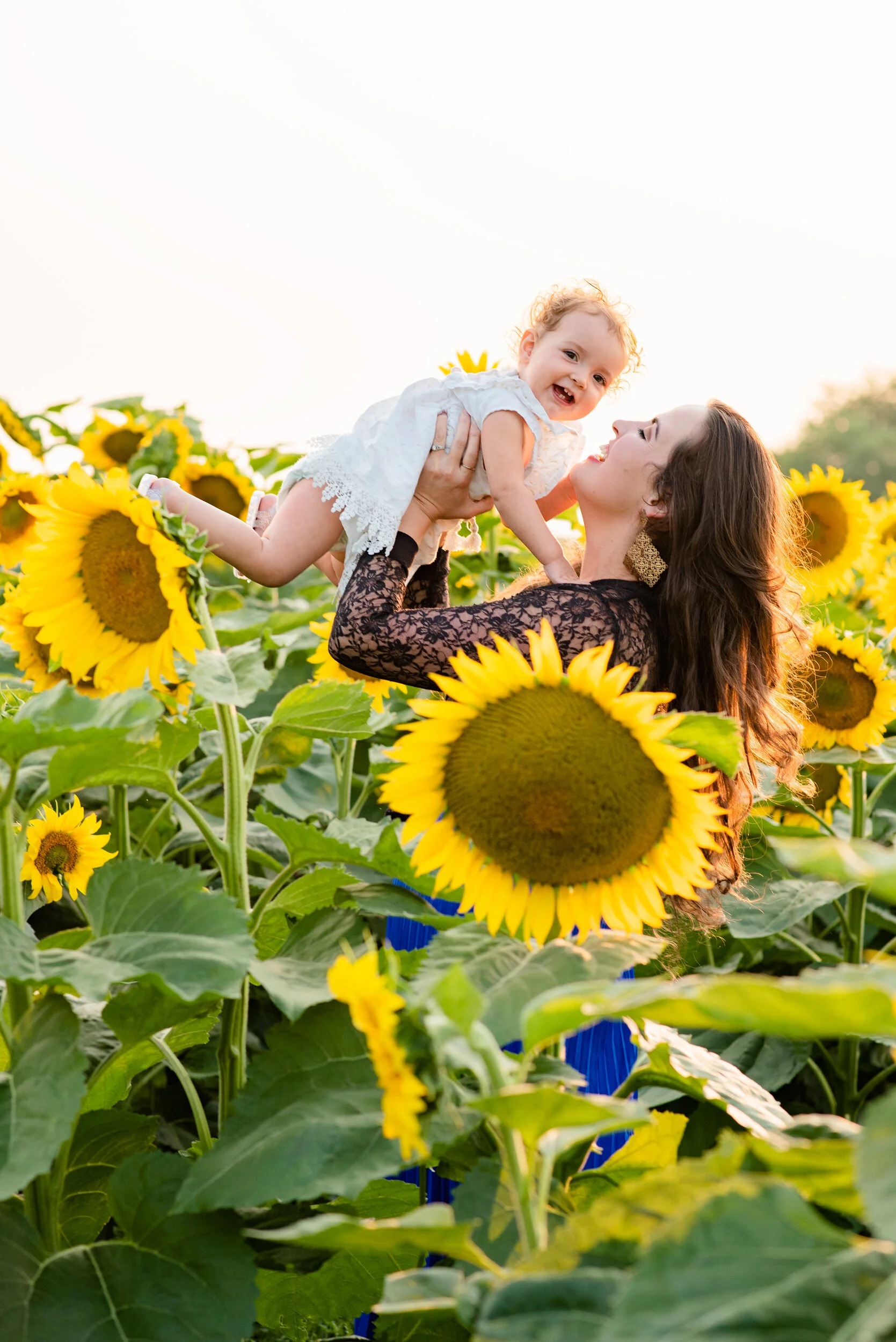 A woman holding a smiling young girl in a sunflower field at sunset.