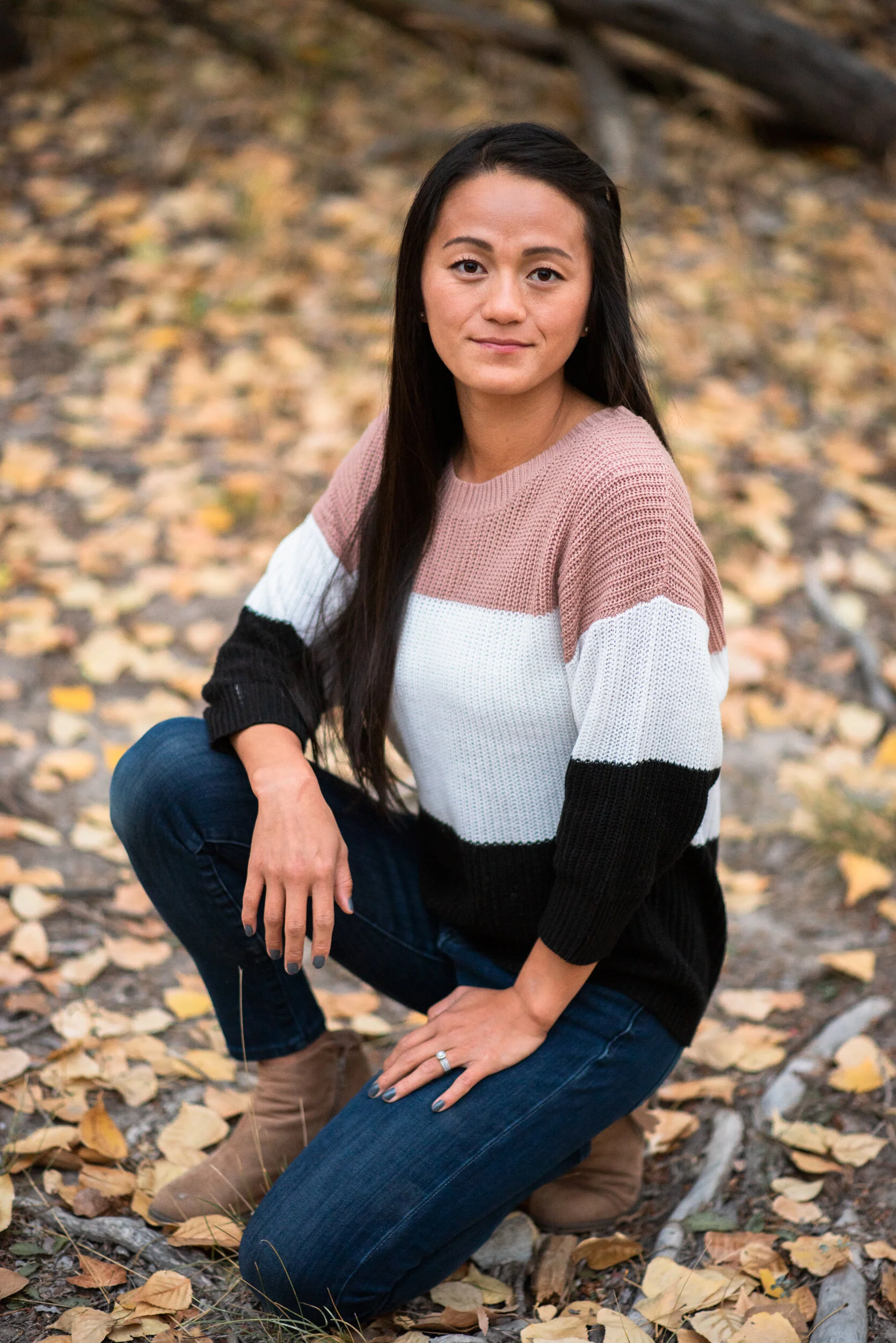 A woman kneeling outdoors on fallen autumn leaves, wearing a pink, black, and white striped sweater, with long dark hair and a neutral expression.
