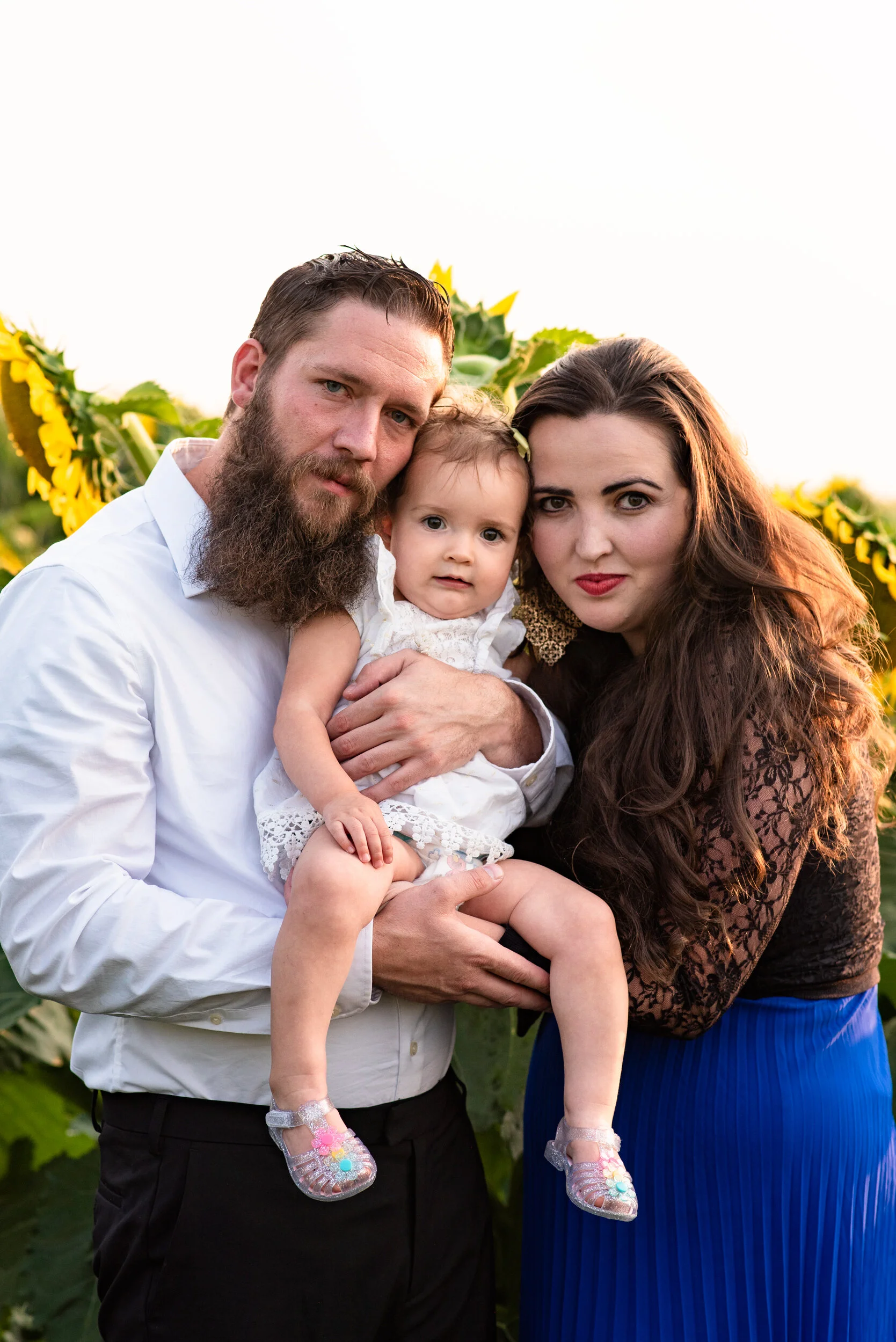 A couple and a young girl posing outdoors among sunflower plants, with the man wearing a white shirt, the woman in a black lace top and blue skirt, and the girl in a white dress with shiny sandals.