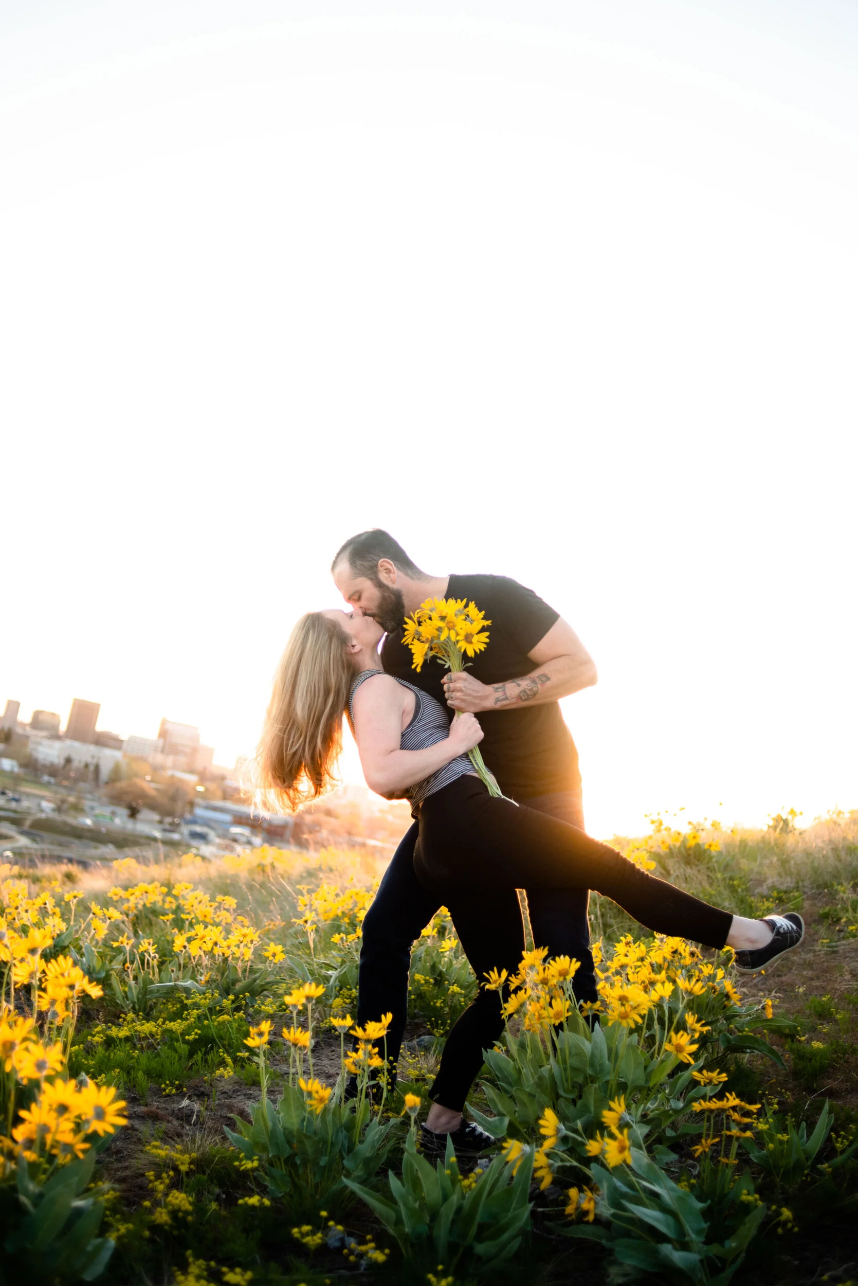 A couple sharing a kiss in a field of yellow flowers at sunset, with city buildings in the background.