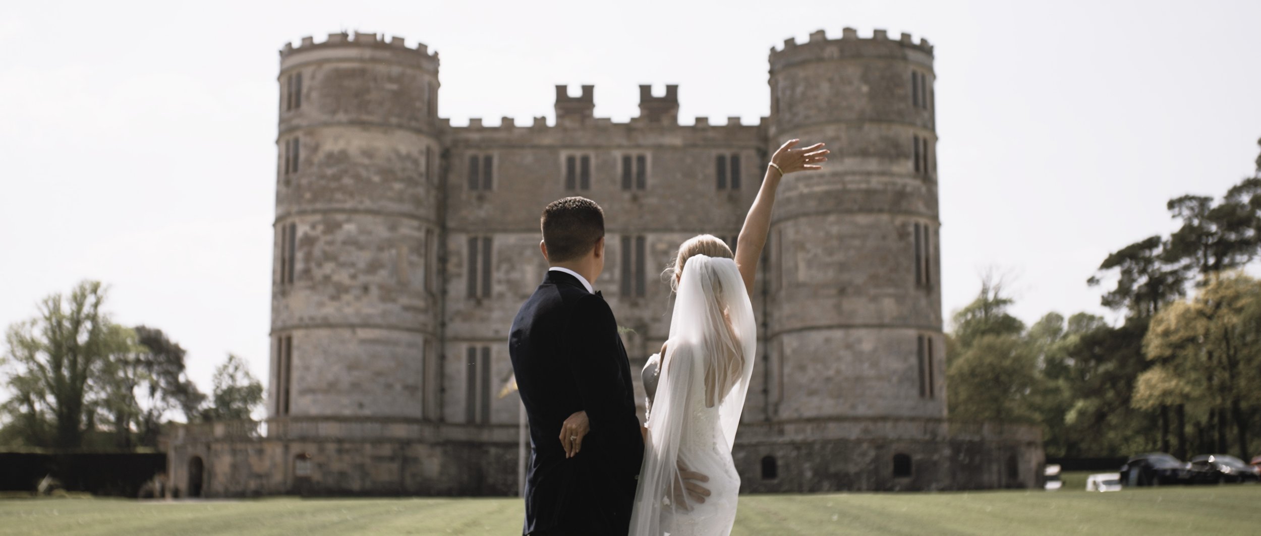 Couple standing in front of Lulworth Castle waving back at wedding guests