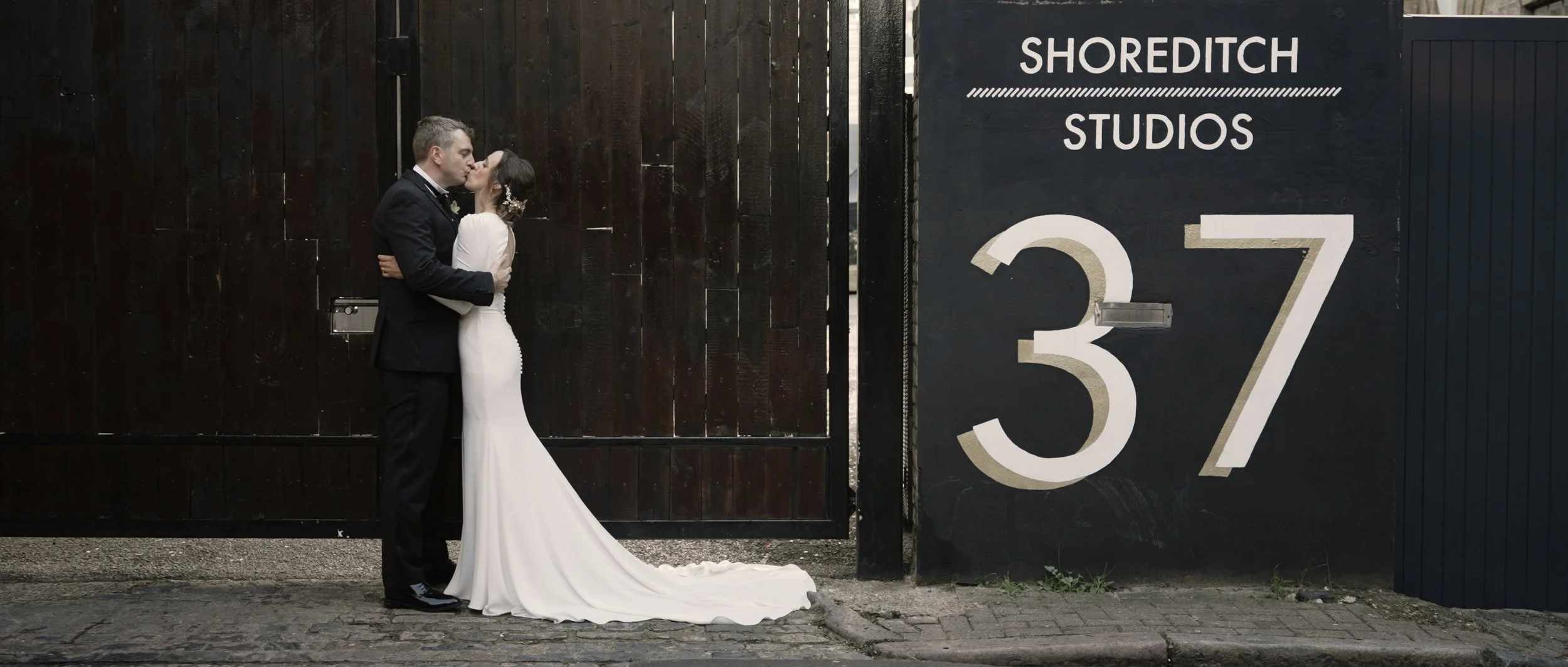 A newlywed couple sharing a kiss outside a black gate and fence, with a sign that reads 'SHOREDITCH STUDIOS 37'.