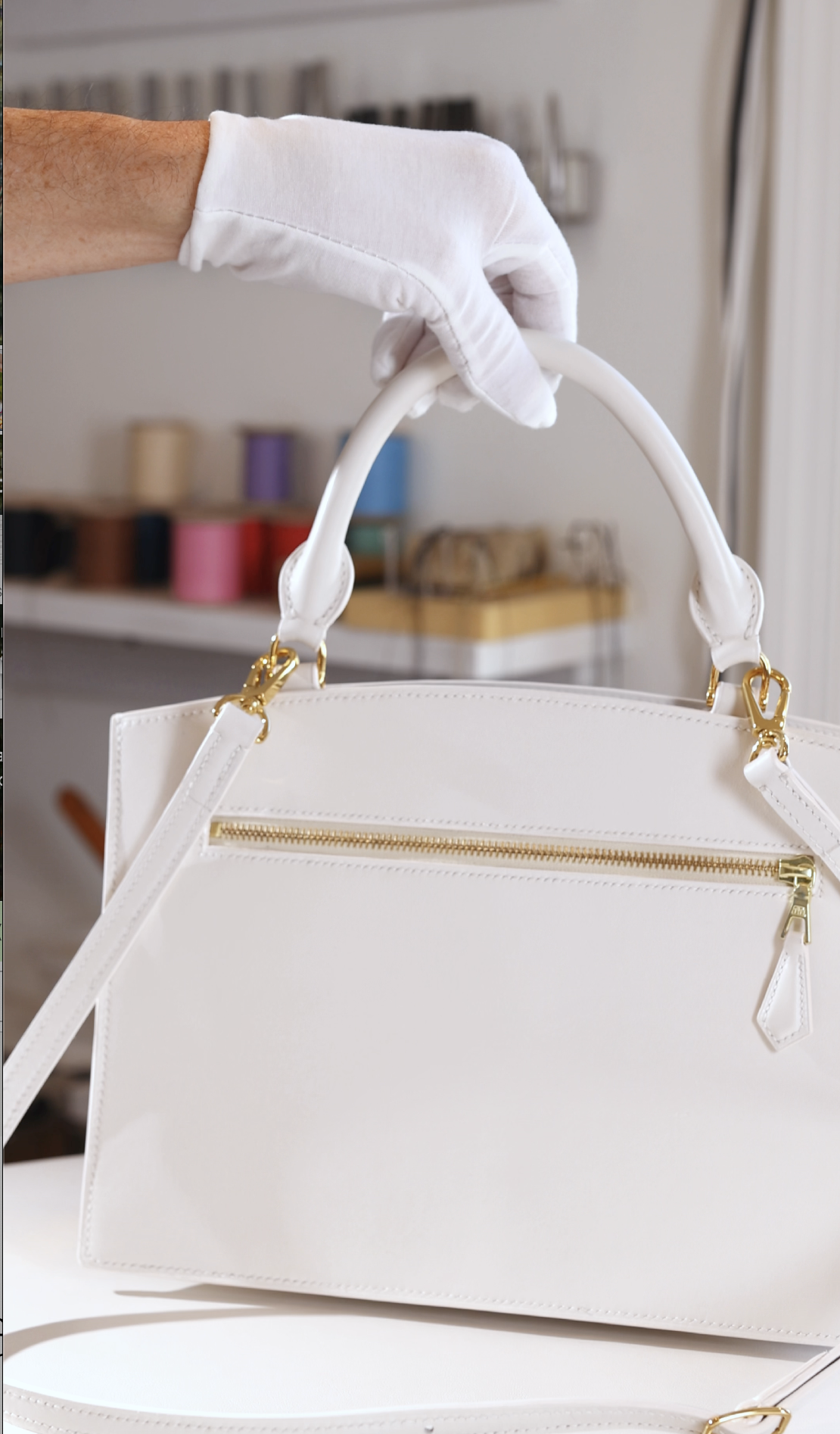 A hand wearing a white glove holding a white handbag with gold zippers and hardware in front of a background with colorful spools of thread and sewing supplies.