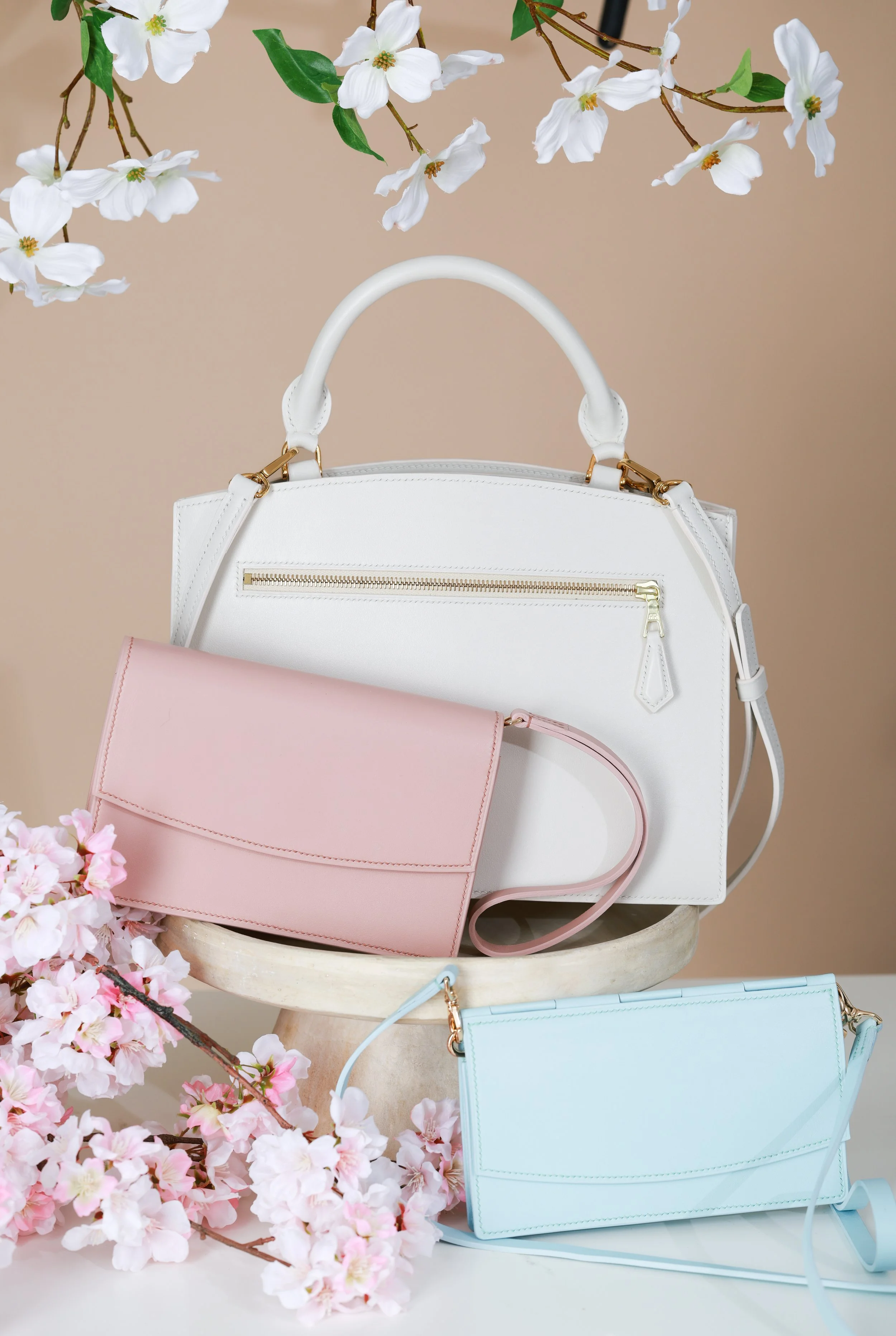 White handbag with gold zipper, pink clutch with wrist strap, and light blue clutch with wrist strap, surrounded by pink cherry blossom flowers and white blossoms on a beige background.