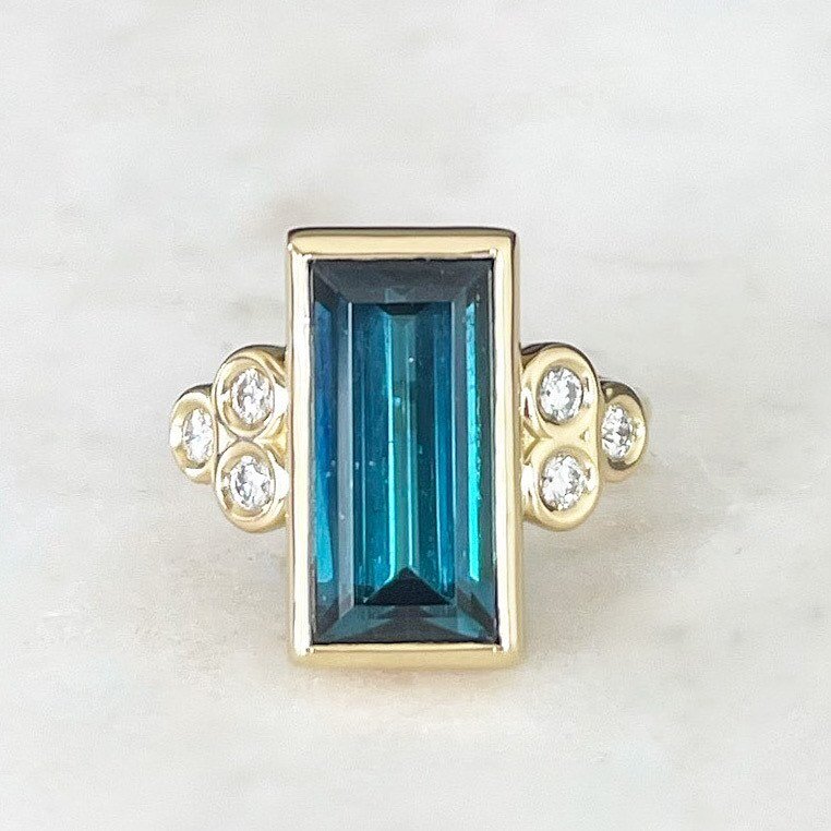 JOLIE CUSTOM RING: ONE-OF-A-KIND INDICOLITE TOURMALINE AND DIAMOND 
Simply delicious. The rustic warmth of  18-karat yellow gold complements the unique radiance of a true indicolite straight baguette cut tourmaline nestled in sparkling white diamonds