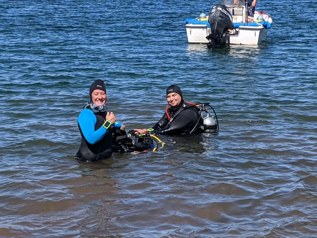 Two scuba divers in wetsuits and diving gear standing in shallow water near a sailboat, smiling at the camera.