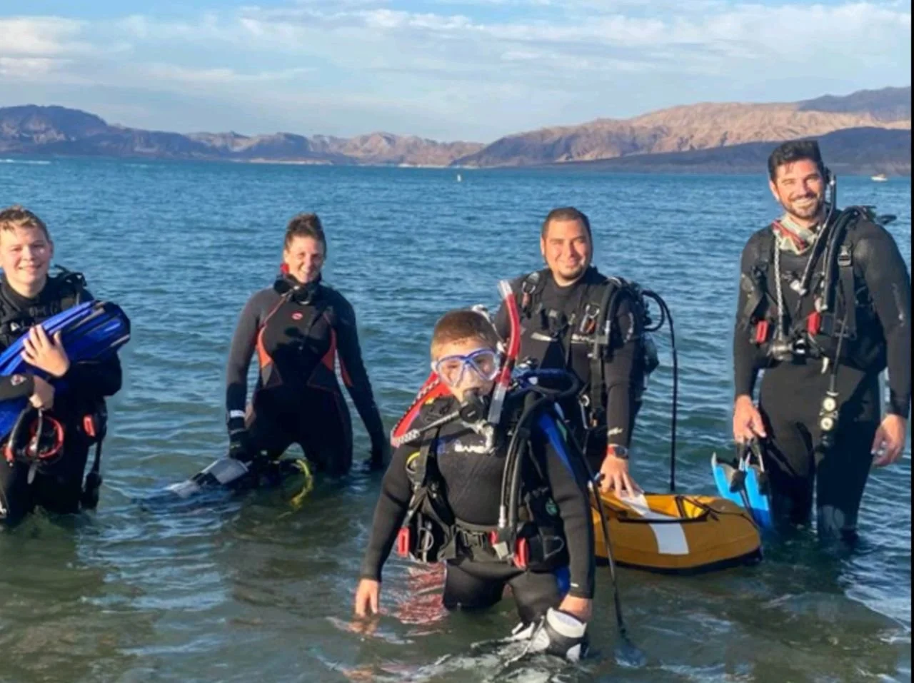 Group of five people in diving gear wading in water at a lake or ocean with mountains in the background.