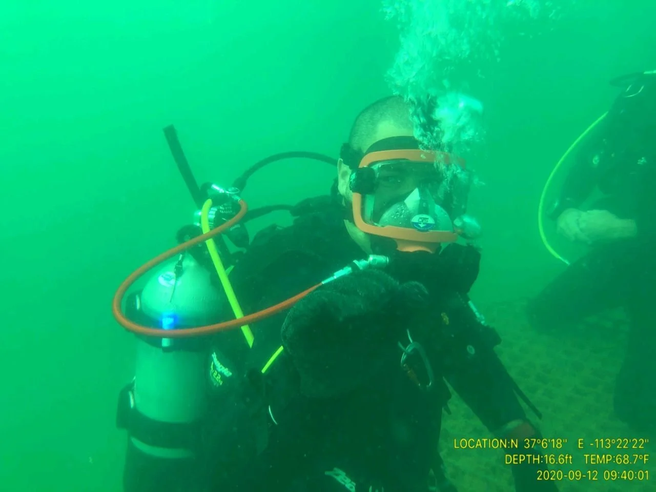 A scuba diver underwater wearing a mask, with bubbles escaping, and a tank on their back, giving a thumbs-up sign. Another diver is partially visible on the right. Green water surrounds them.