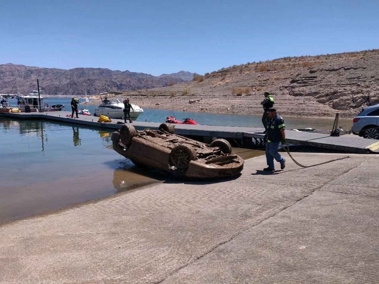 An underwater vehicle overturned on the boat ramp, with tow workers on shore pulling it out. Several boats are docked at the marina, and mountains are visible in the background under a clear blue sky.
