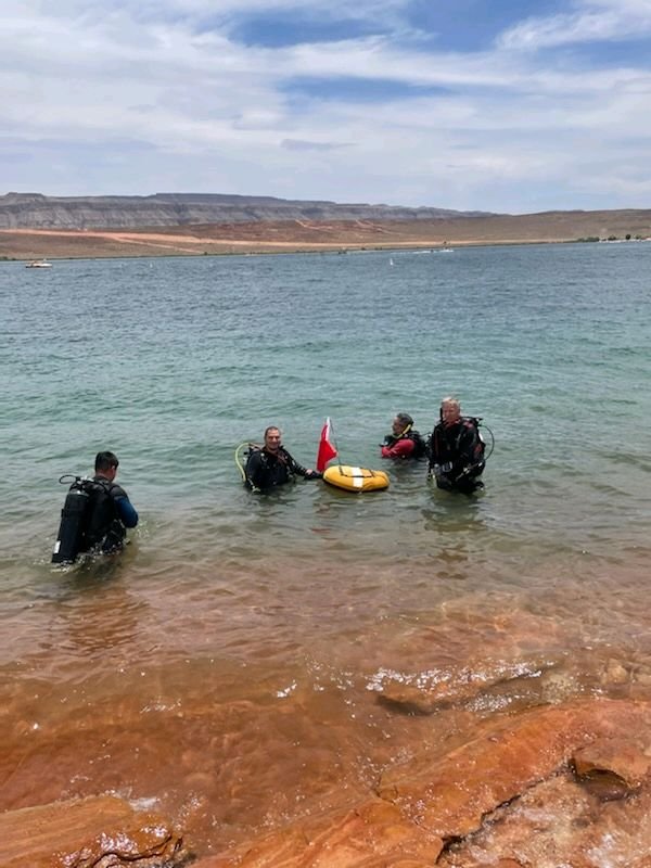 Four scuba divers in a lake, with two holding a yellow flotation device with a Canadian flag, at the shore with a distant desert landscape and cloudy sky.