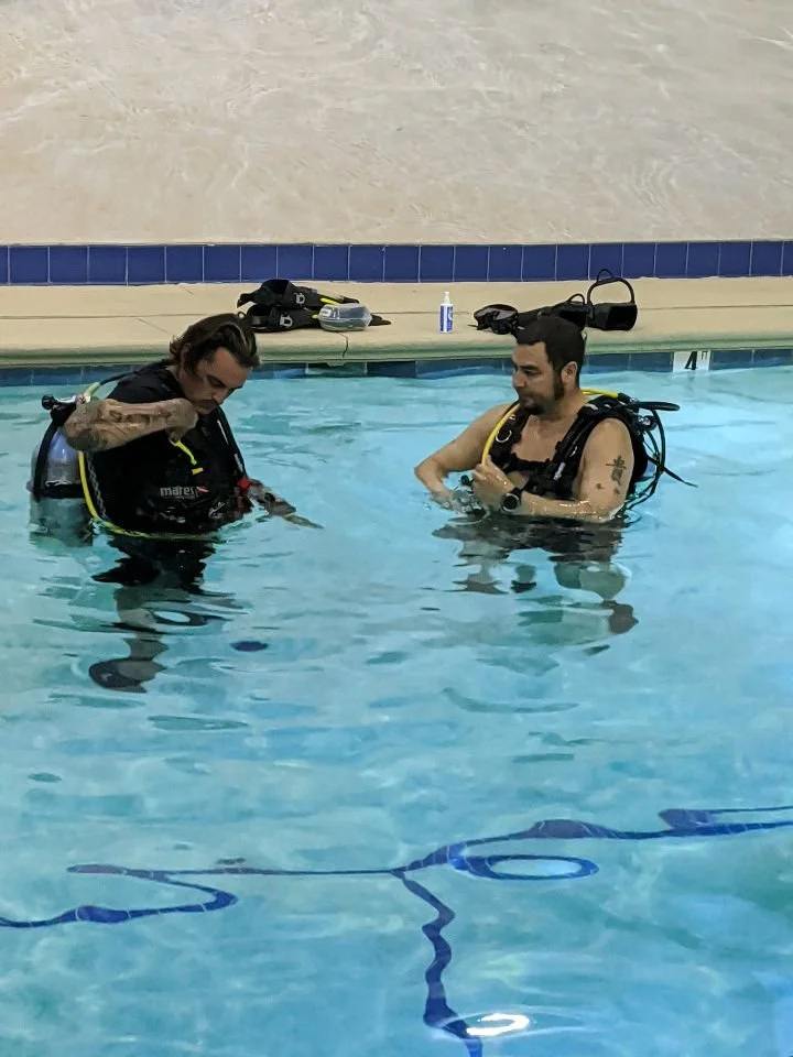 Two men in a swimming pool wearing scuba diving gear, preparing for a scuba lesson.