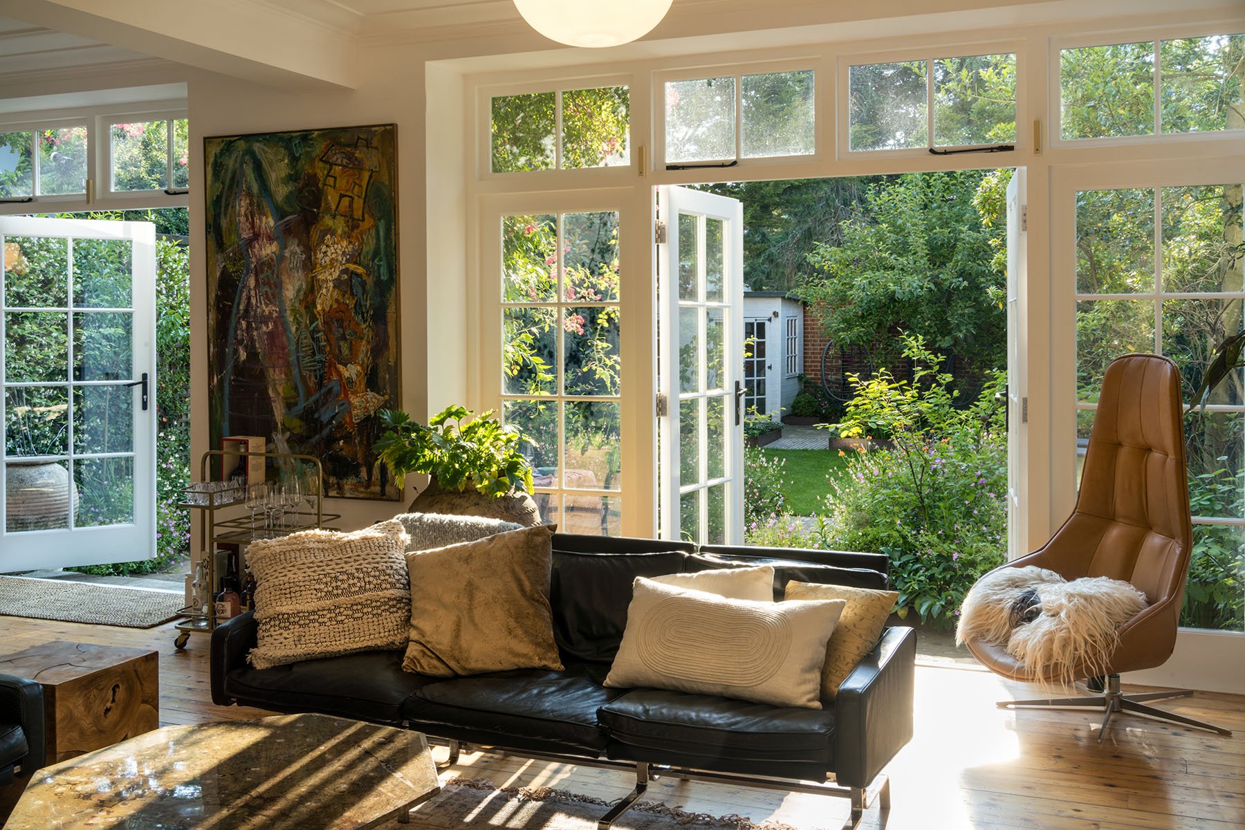 Bright living room with large open windows, black leather sofa with pillows, tan modern chair with a dog on a sheepskin rug, and a view of a lush garden outside.