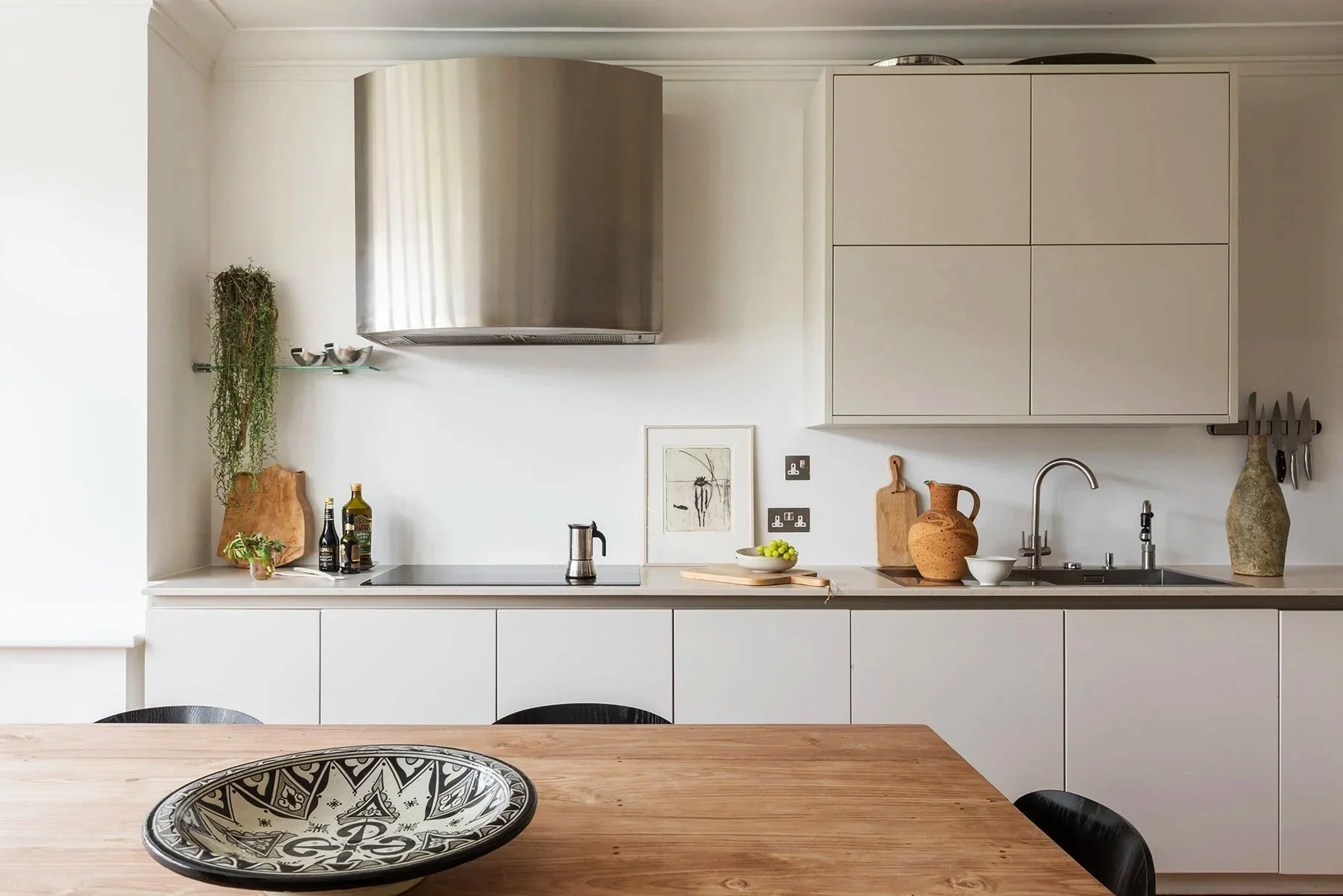 Modern minimalist kitchen with white cabinets, stainless steel range hood, wooden table, decorative pottery, and plants.