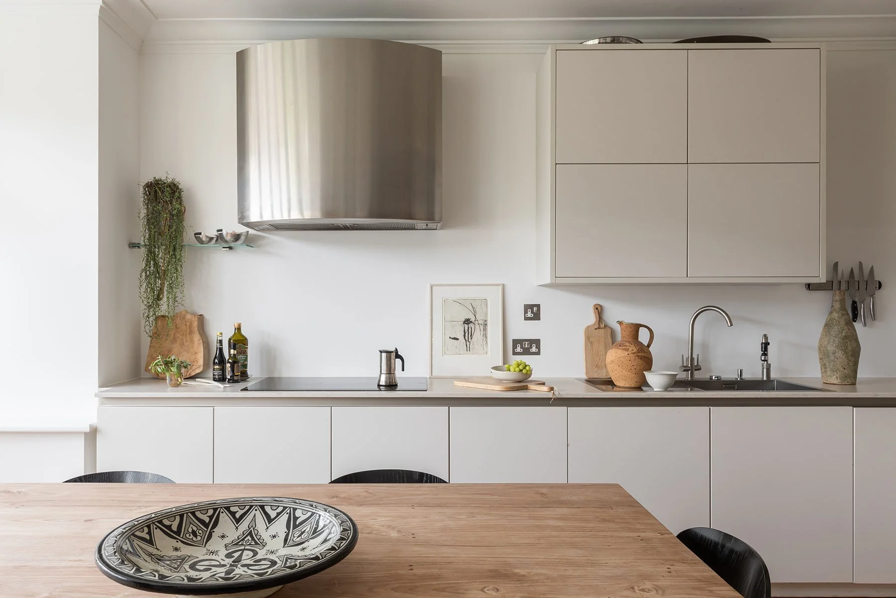 Modern minimalist kitchen with white cabinets, wooden table, decorative vases, and potted plants.