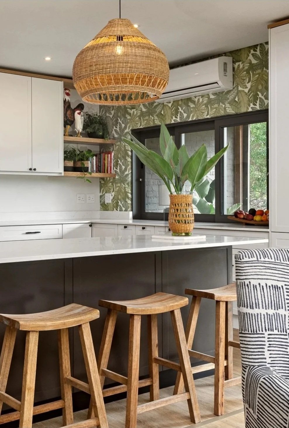 A classic kitchen in Loxley Estate, Ballito featuring a white countertop, black kitchen island, three wooden stools, a large green plant in a woven basket, a window with greenery outside, and decorative wallpaper with a leafy pattern.