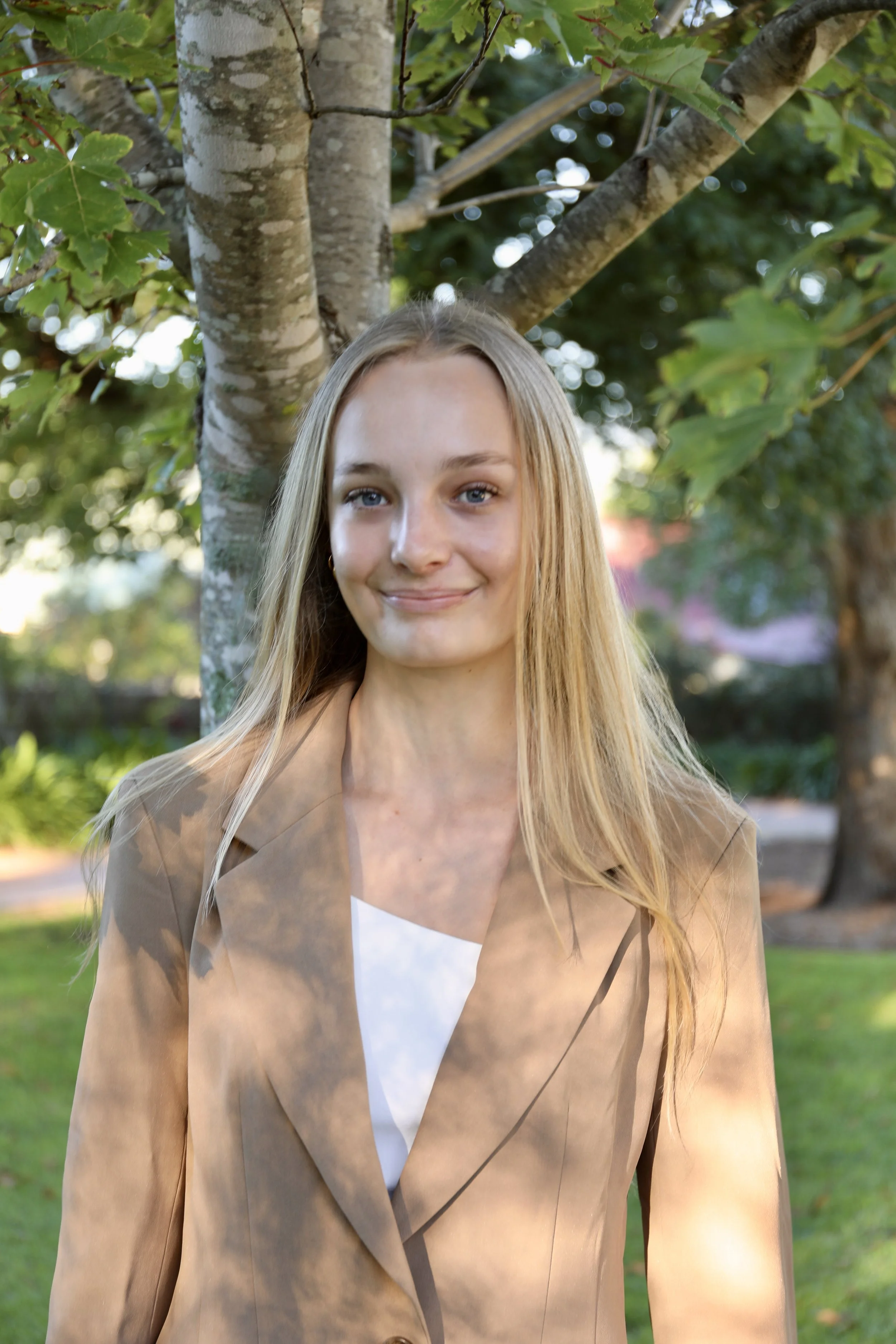 A young woman with long blonde hair, smiling, standing outdoors near a tree with green leaves.