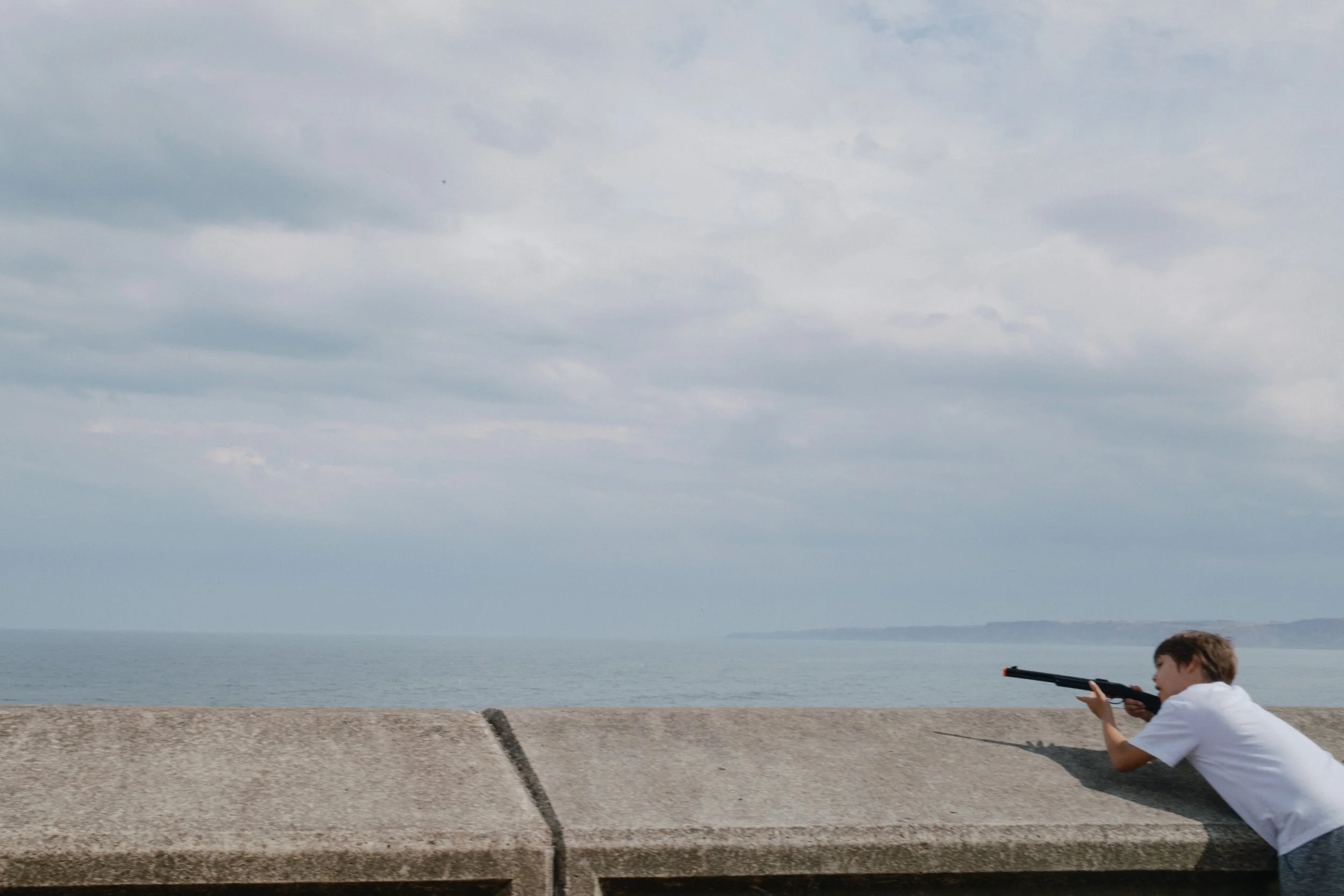 A young boy in a white shirt crouches by a concrete barrier, aiming a toy gun towards the sky by the seaside during the day with cloudy weather.