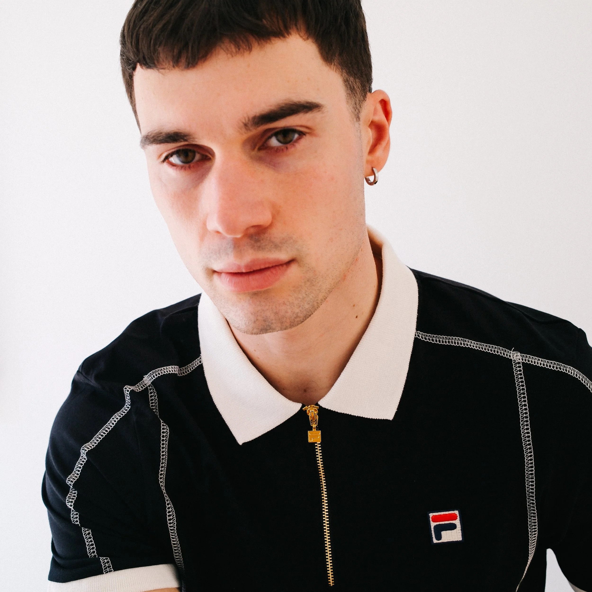 Close-up of a young man's face with short brown hair, earrings, and wearing a black and white jacket with a zipper and brand logo, against a plain white background.